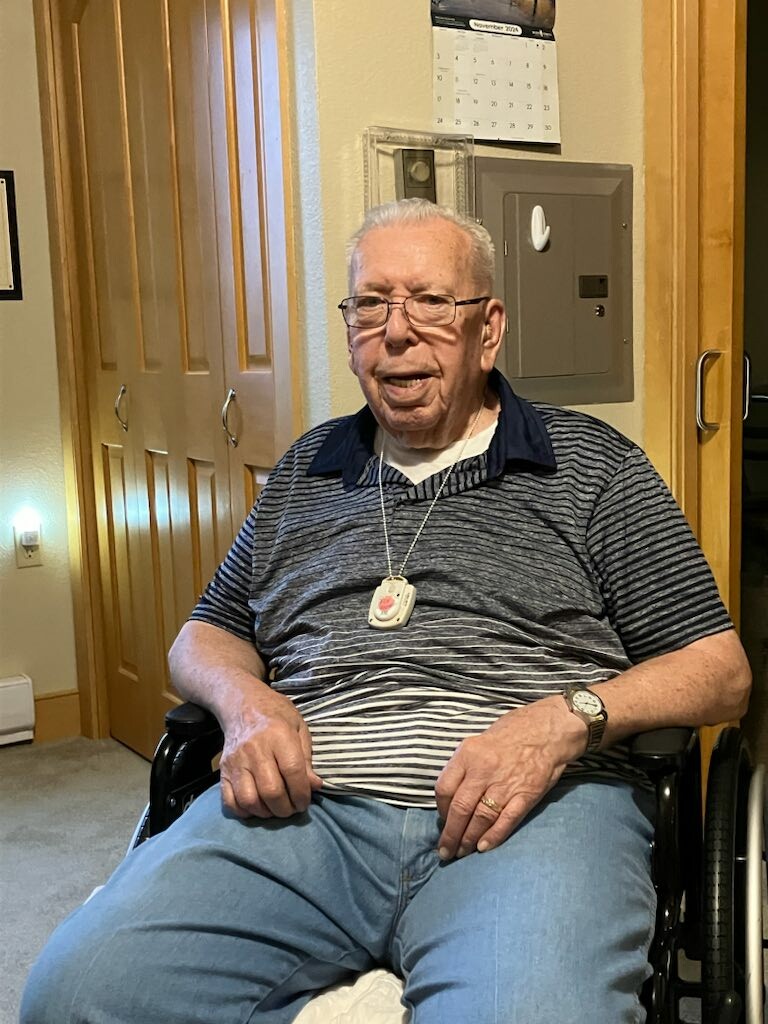 Older man in a wheelchair indoors, wearing a blue striped polo and jeans, smiling at the camera.