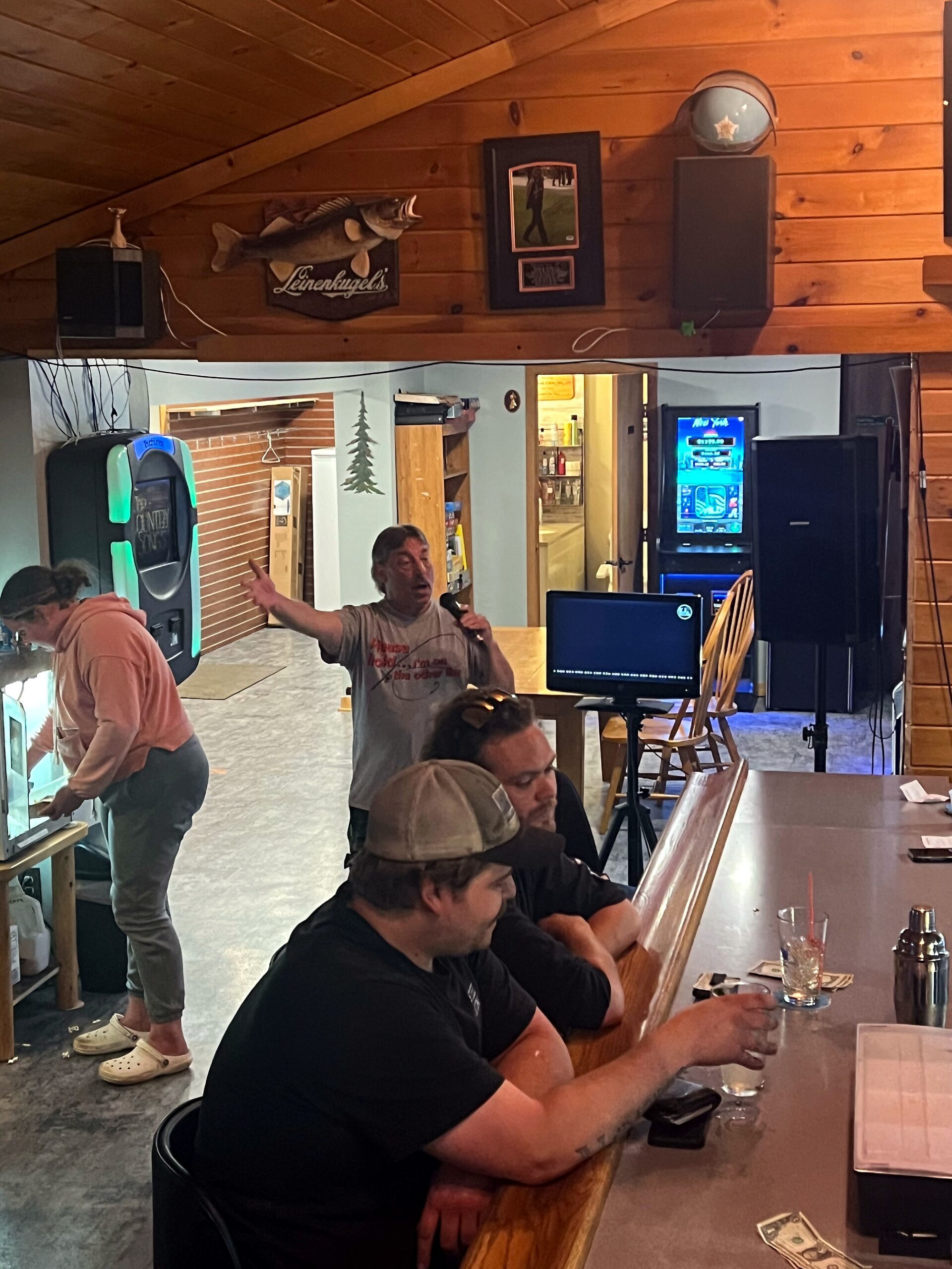 Man with a microphone singing karaoke at a bar counter as other patrons sit nearby in a wood-paneled room.