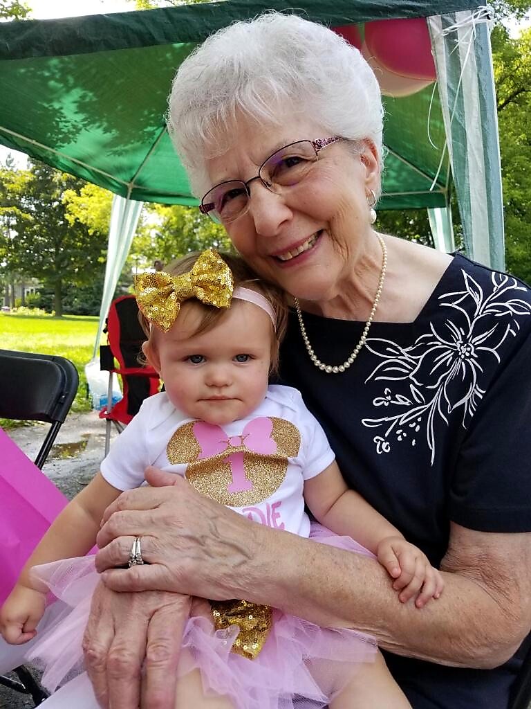 Smiling grandmother with white hair and glasses holds a baby in a pink tutu and gold bow at an outdoor party under a green canopy.