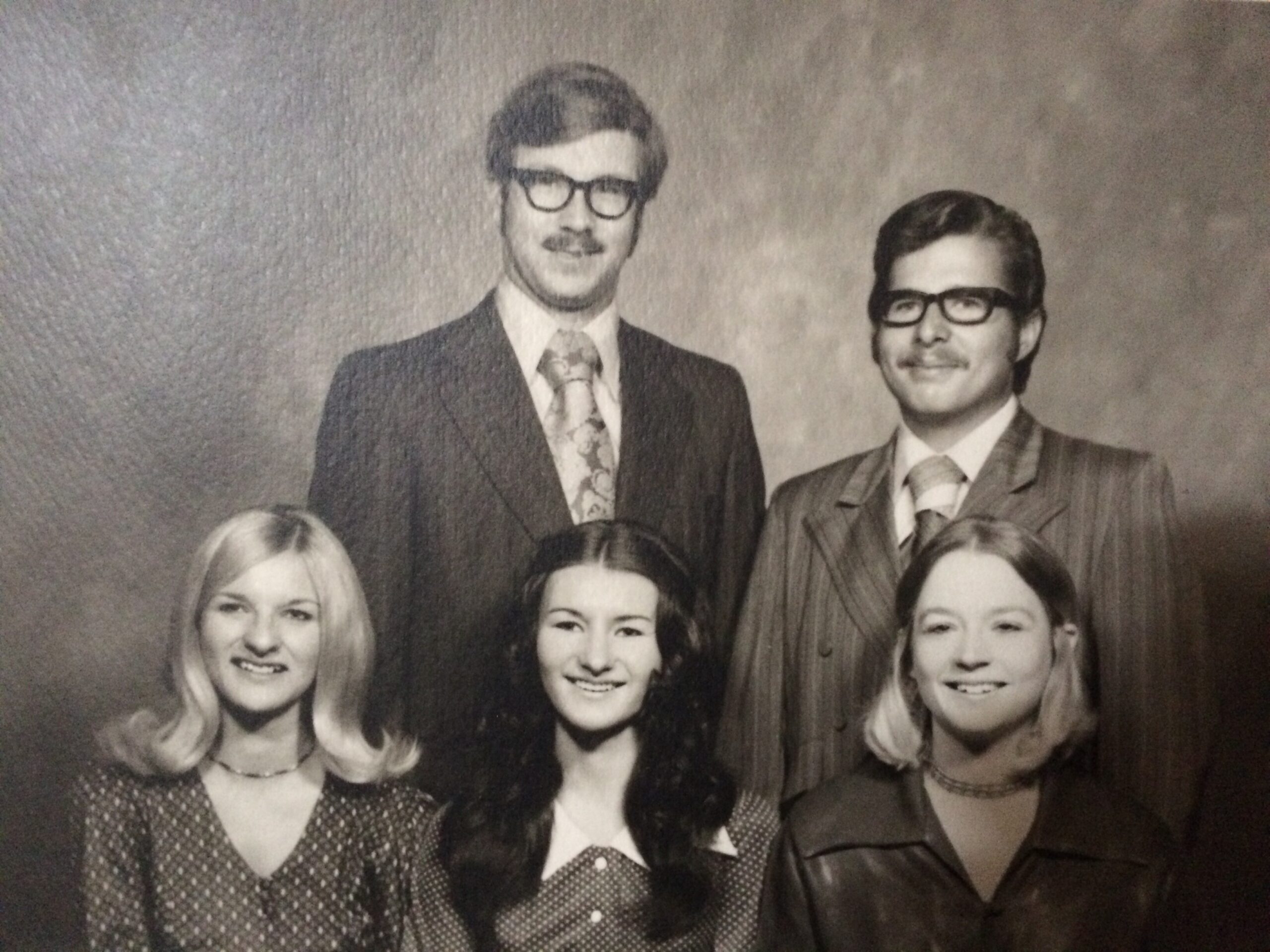 Black-and-white group portrait of adults in 1960s-style formal attire, smiling for a studio photo.