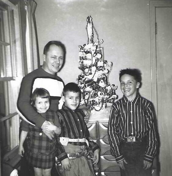 A smiling man with three children posing by a small Christmas tree indoors in a living room.