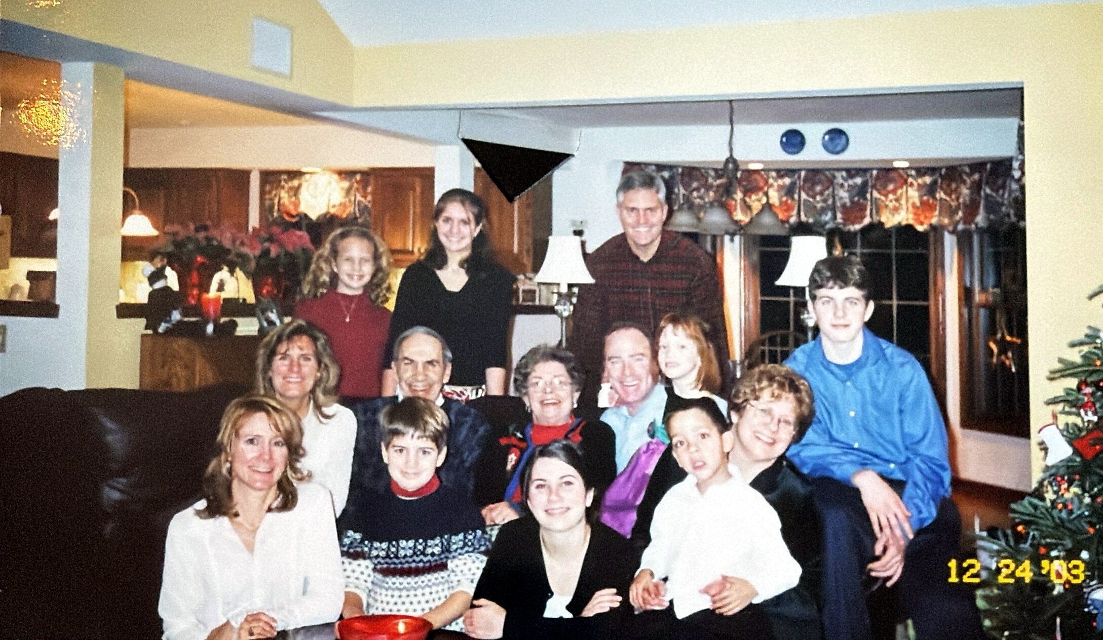 Family and friends posing for a holiday photo in a living room, with a Christmas tree and festive decor in the background (12/24/03).**Note**: Include date if relevant to context.