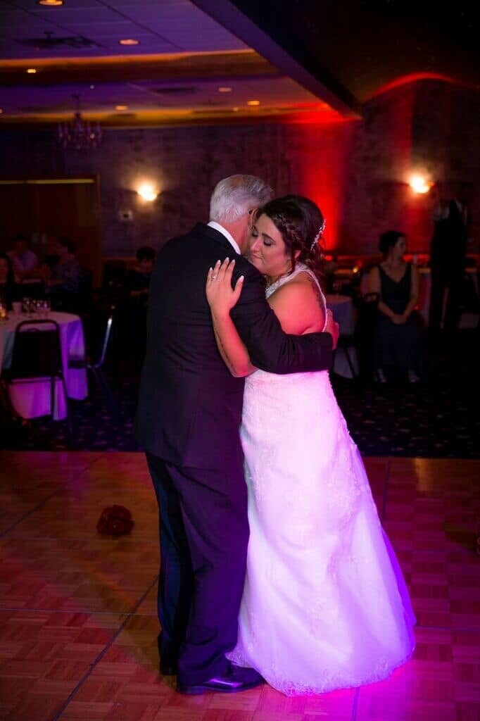 Bride in a white gown dancing with an older man in a dark suit on a lit dance floor at a wedding reception, colorful lighting in the background