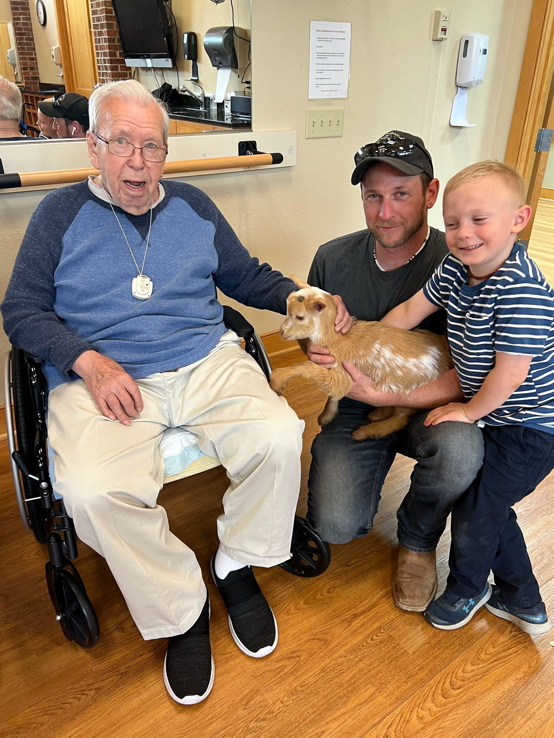Elderly man in a wheelchair, wearing a blue sweater, smiling at a small goat held by a man and a young boy beside him in a room with wood floors.
