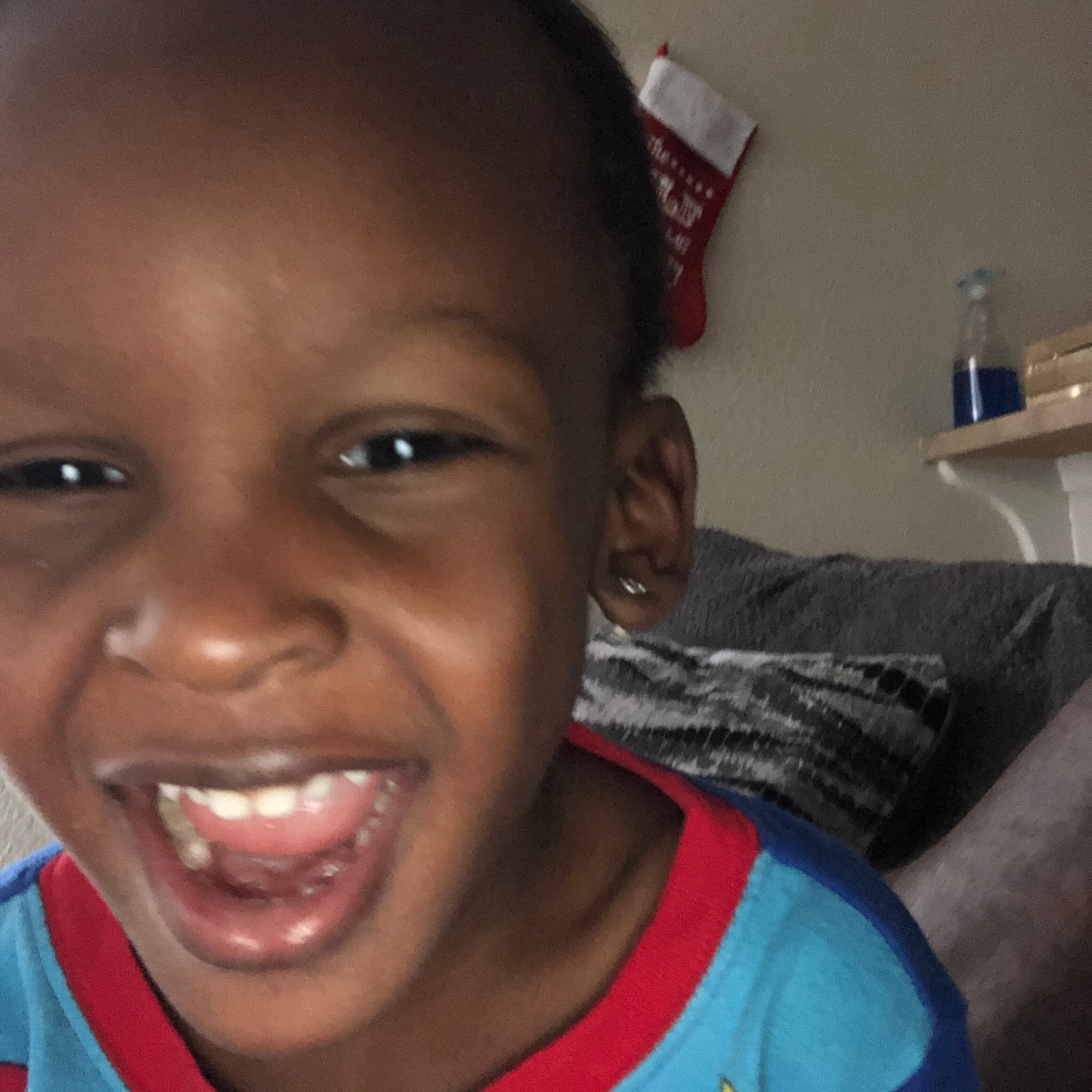 Close-up of a smiling young child with missing front teeth, wearing a blue shirt with red trim in an indoor setting.