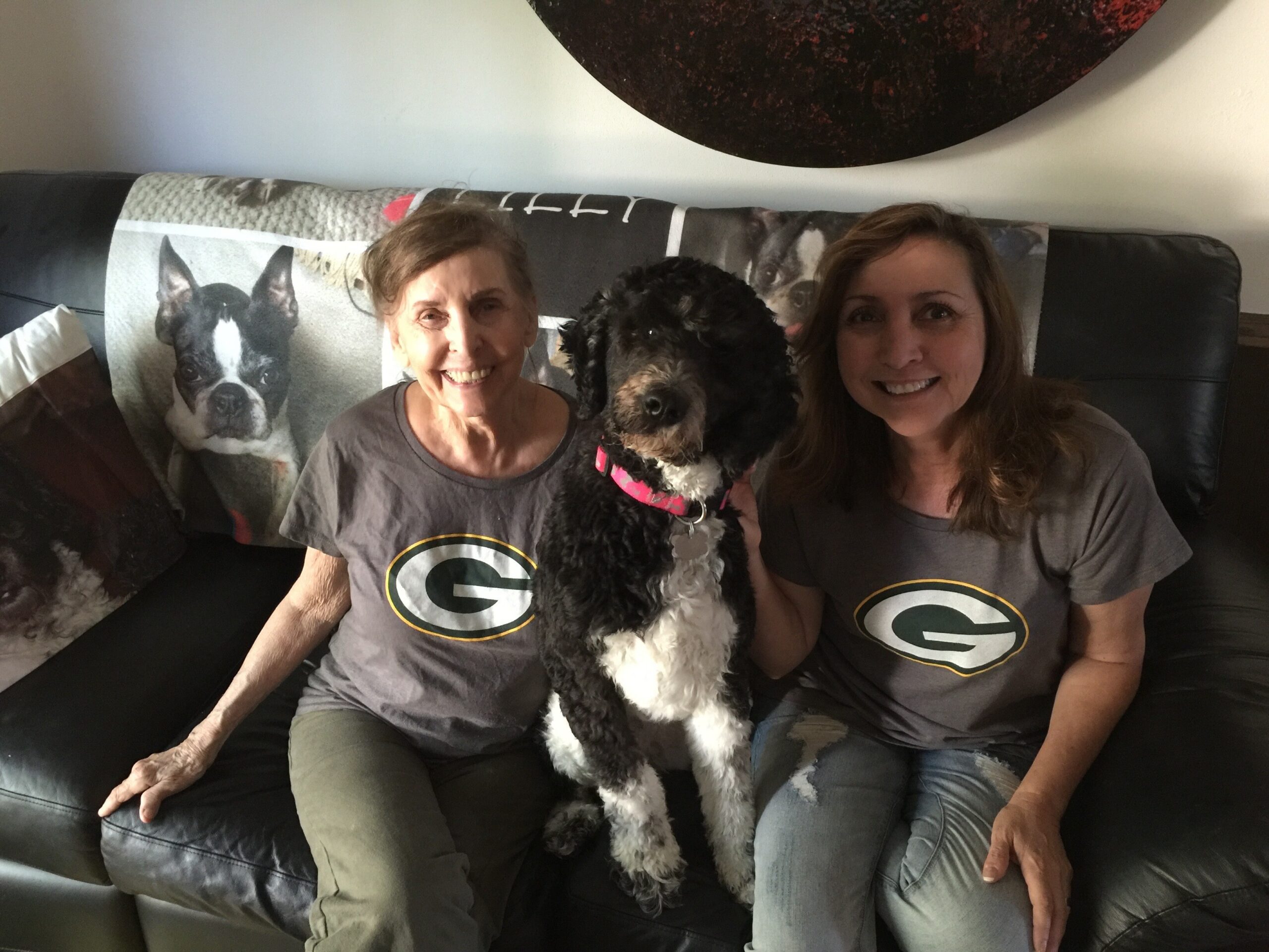 Two women sit on a black leather couch with a black-and-white dog between them, all smiling; they wear gray shirts with a green and yellow Packers logo, and the dog has a pink collar.