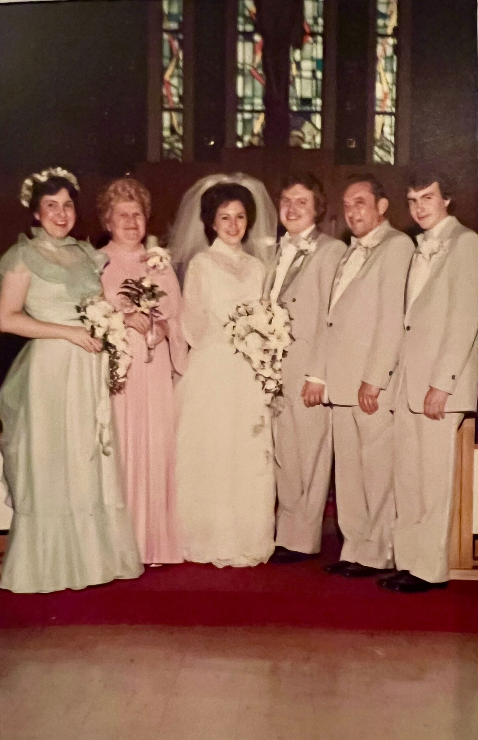 Bride in white gown and veil standing with bridesmaids and groomsmen inside a church with stained glass behind them, posing for a wedding photo.