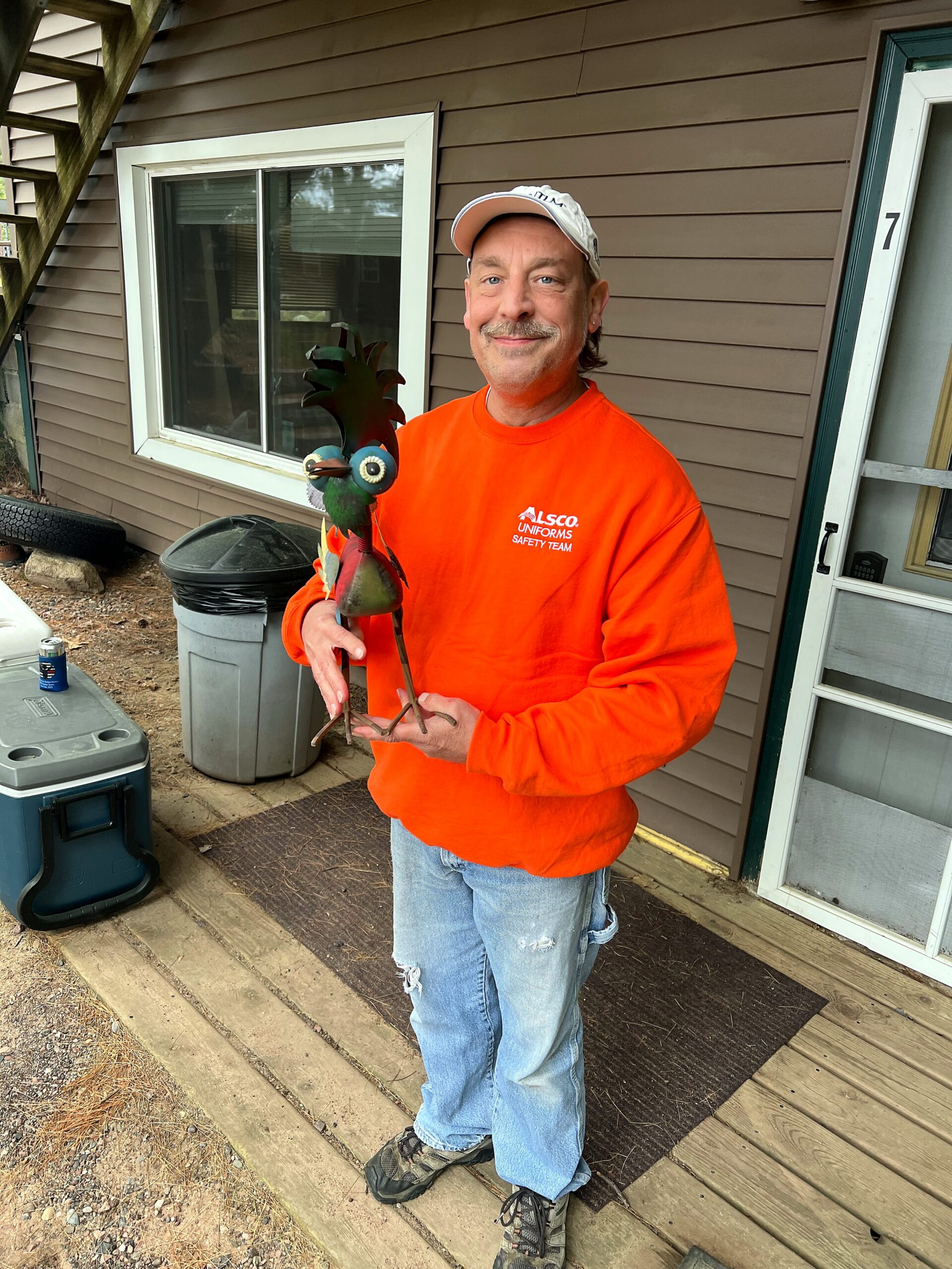Smiling man in an orange shirt holding a metal rooster sculpture outdoors on a wooden porch.