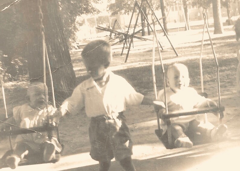Three young children play on swings in a sunny park; one stands nearby while two sit in swings, vintage photo. Silhouetted play scene from childhood.