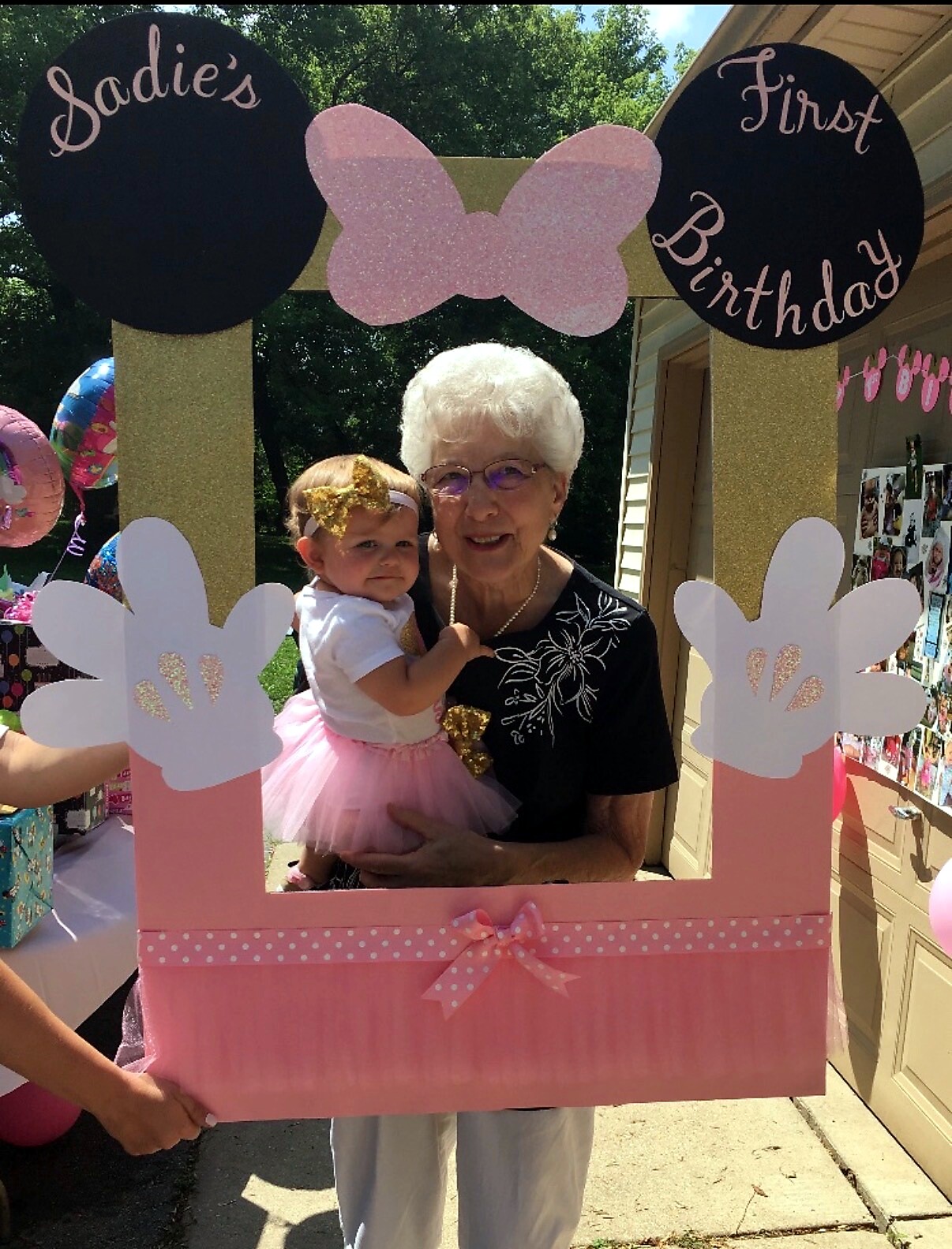 Grandmother holding a baby girl in a pink birthday photo frame reading 'Sadie's First Birthday' outdoors.