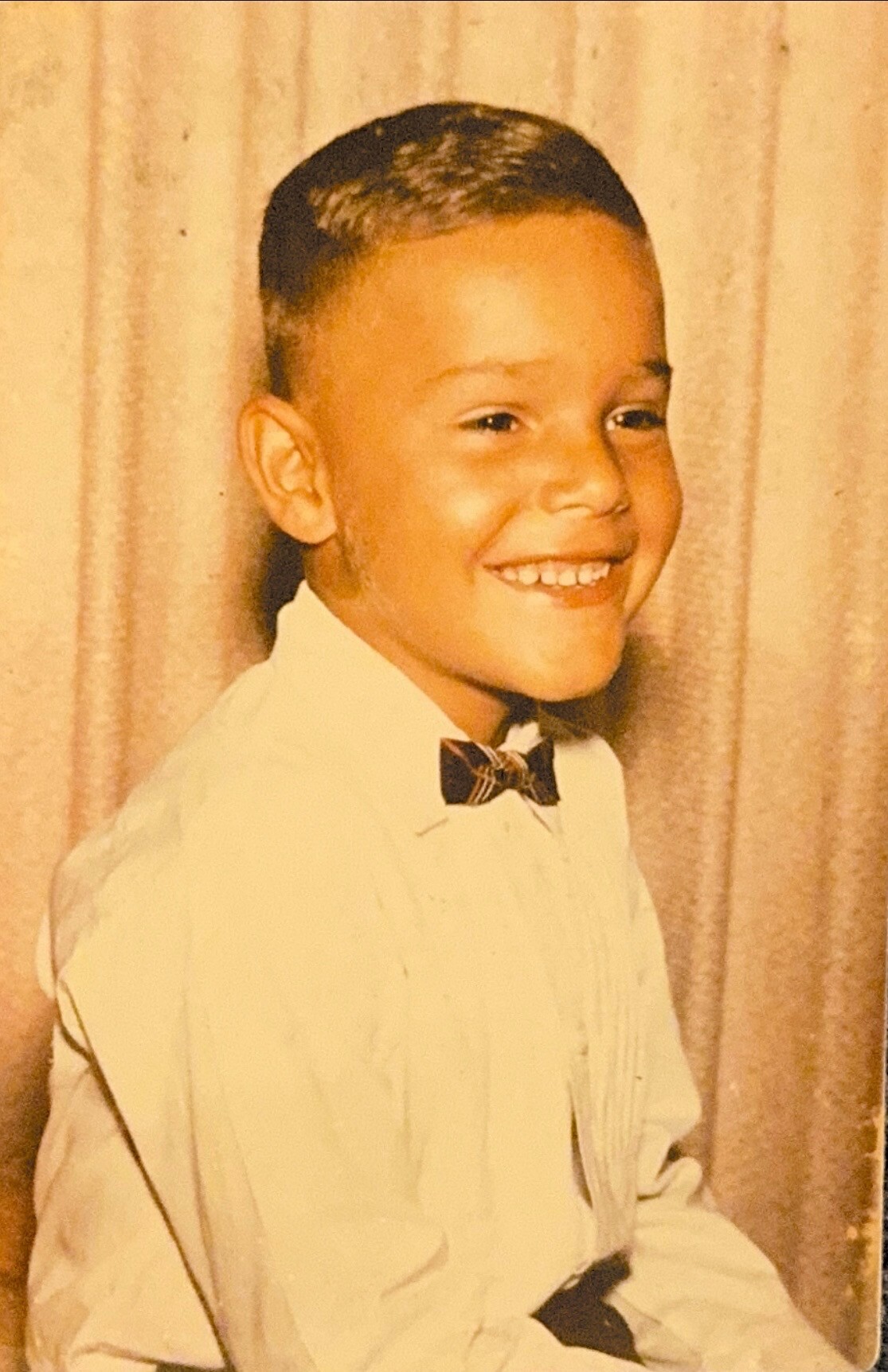 Smiling young boy in a white shirt and bow tie posing in front of a beige curtain.