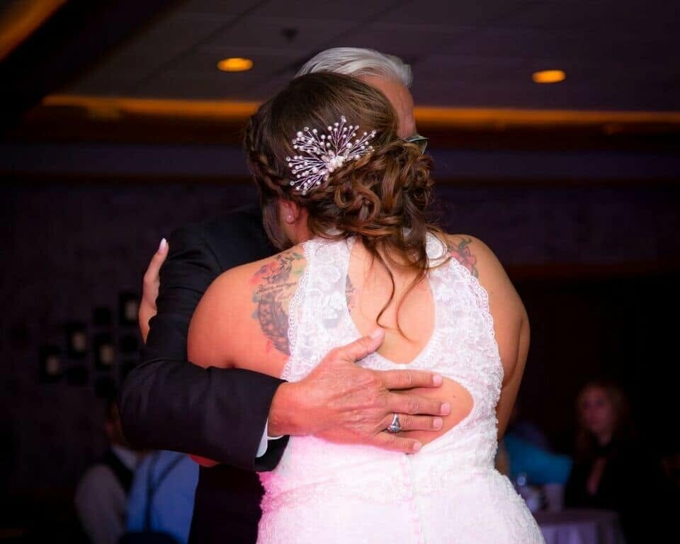 Bride in a white lace gown hugging a man in a dark suit on the dance floor; her hair is styled in an updo with a crystal hair accessory visible from behind.