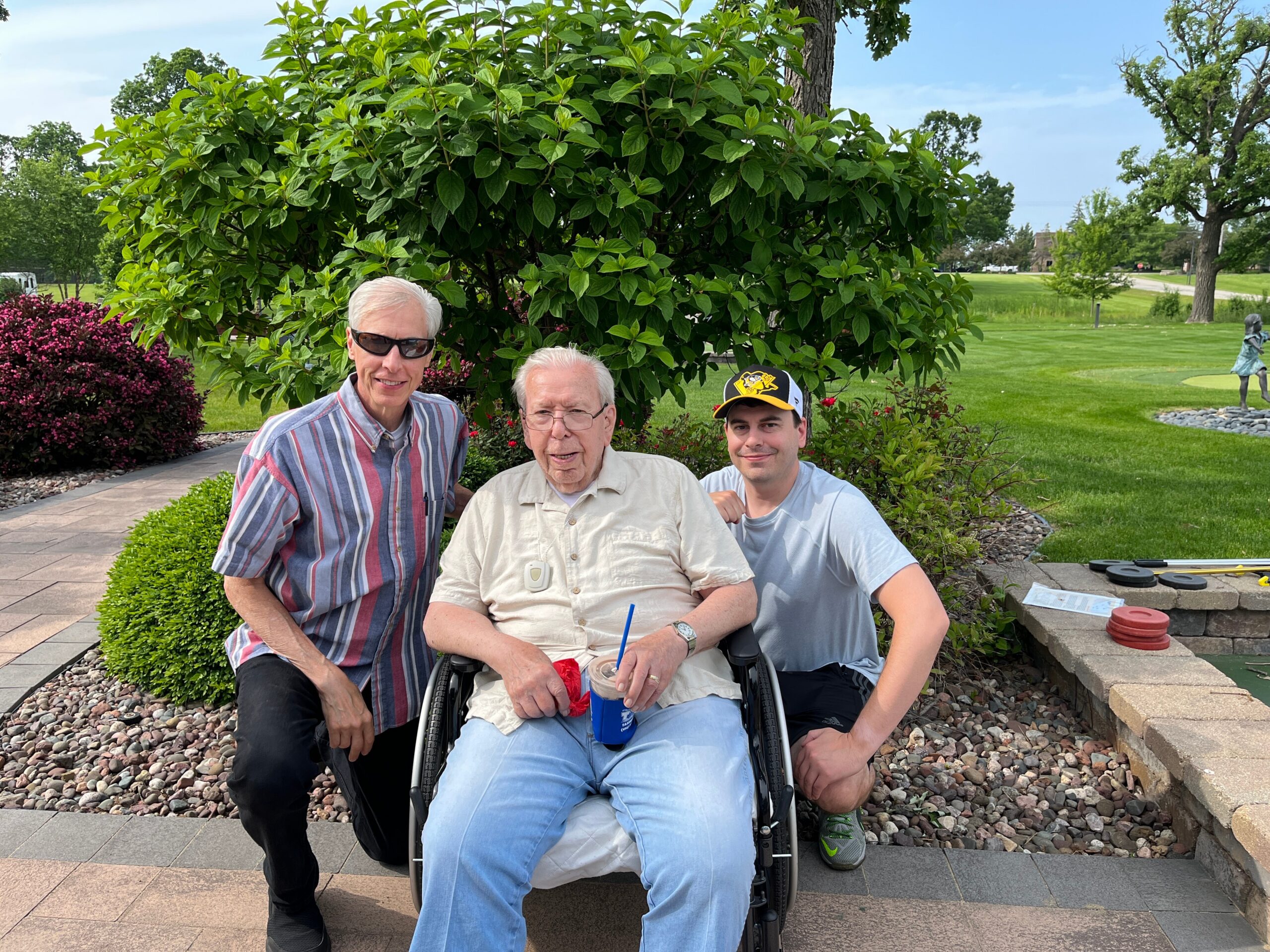 Three men outdoors in a park; one in a wheelchair center foreground, flanked by two standing men, all smiling at the camera.