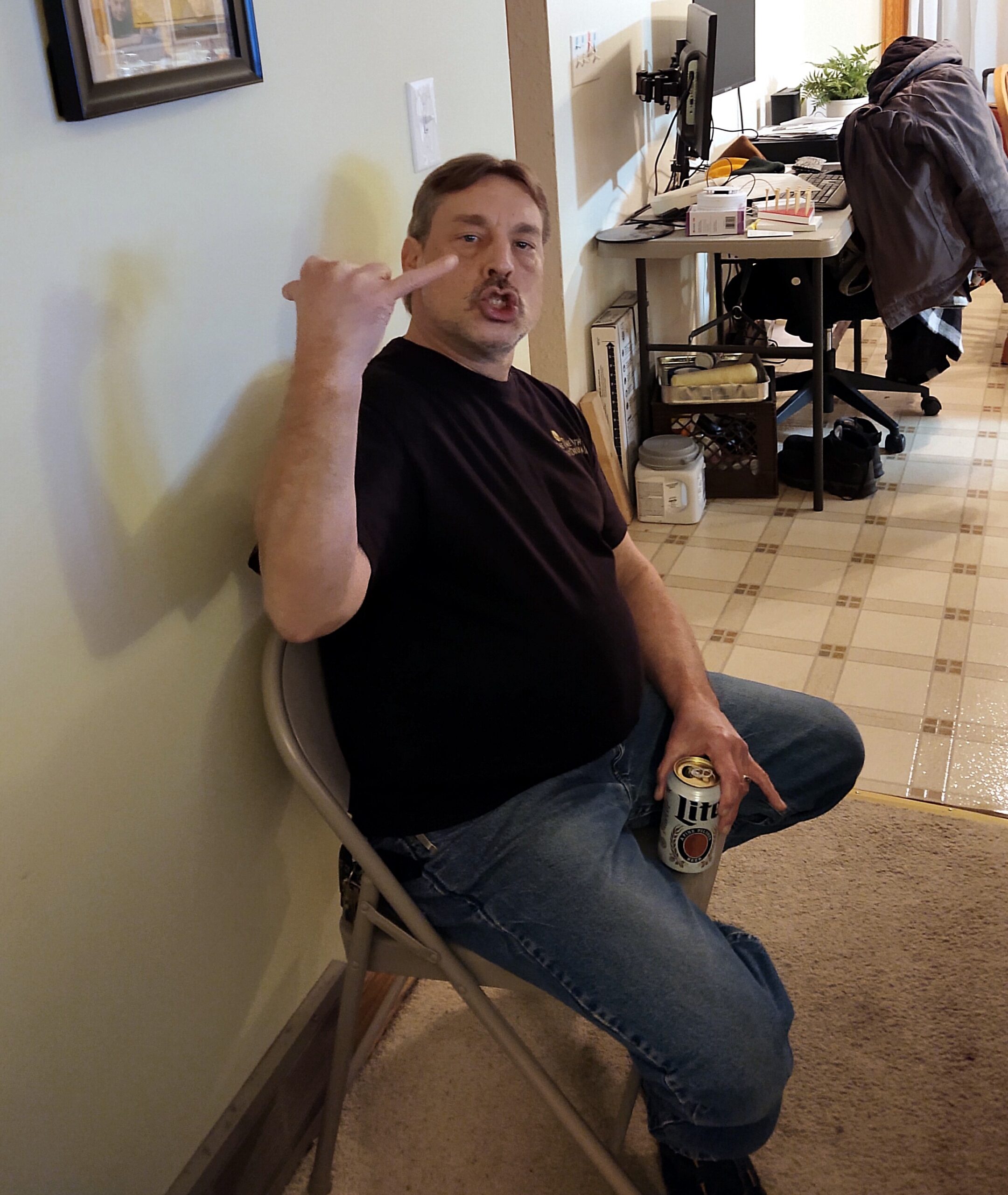 Man seated on a folding chair against a wall, making a rock‑on gesture with a Miller Lite can in his hand.