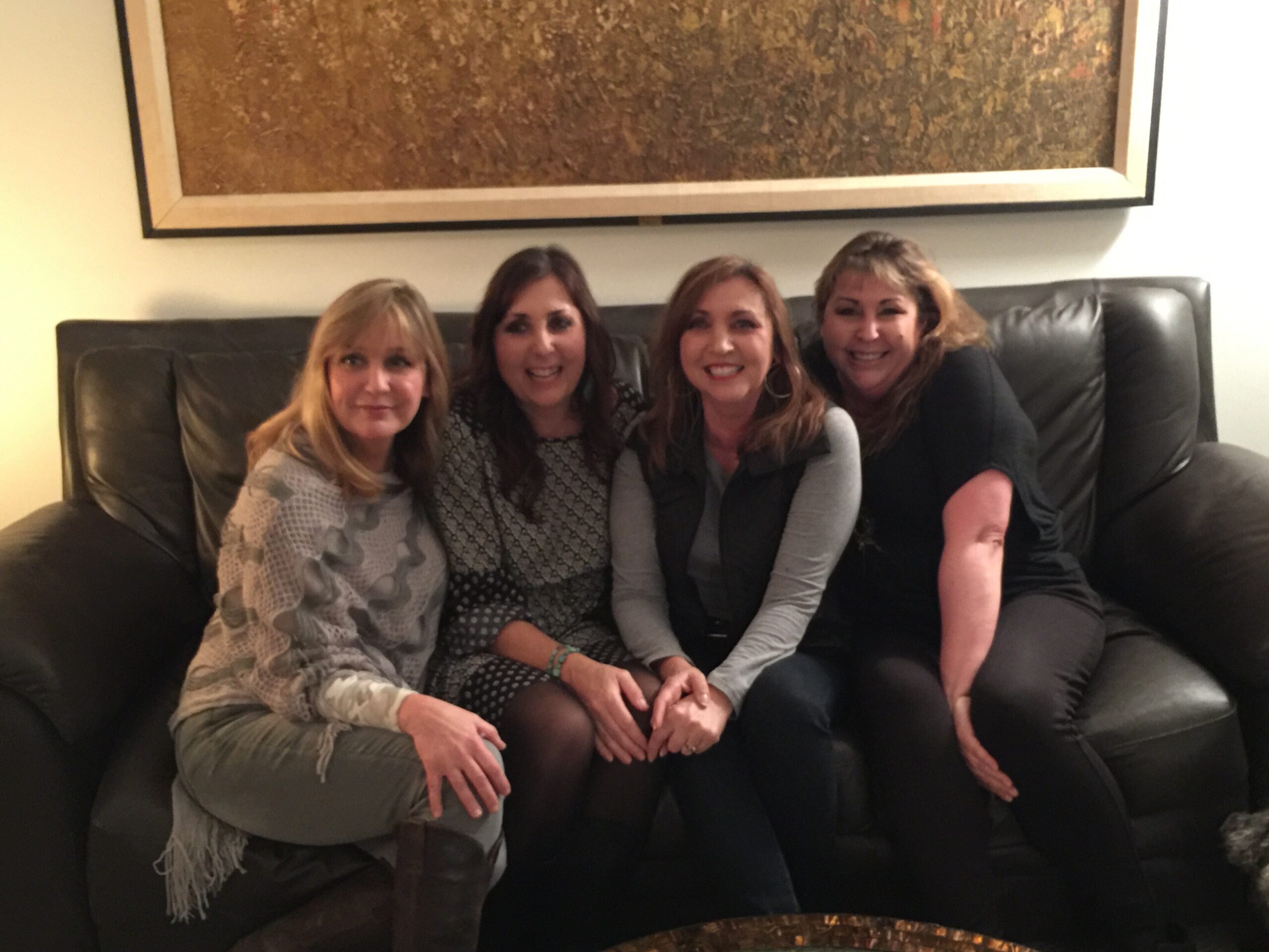 Four women sit close together on a black leather couch, smiling at the camera in a living room with a large framed painting above.