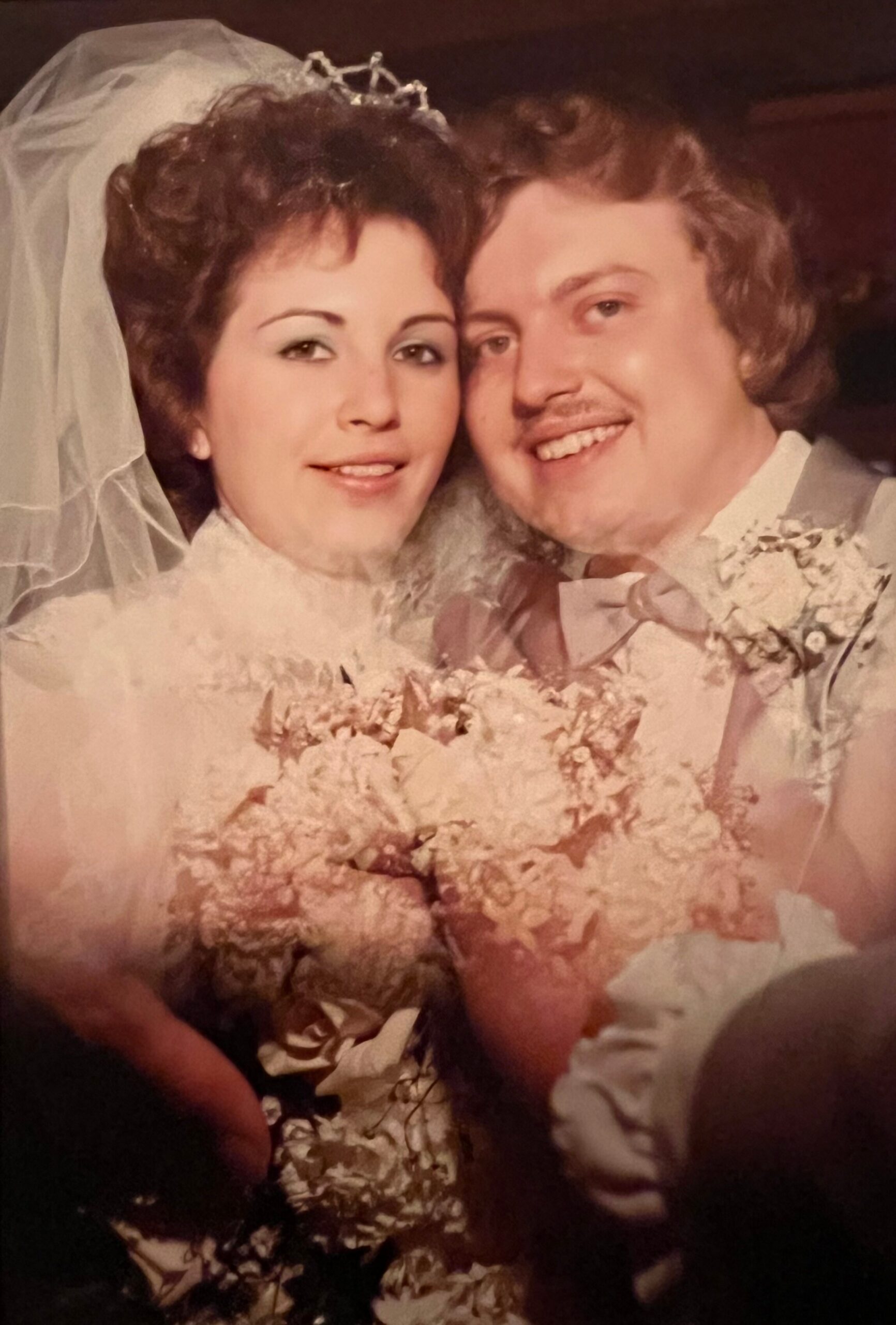 Bride in a wedding gown with veil and bouquet, smiling beside her groom in a suit and bow tie.