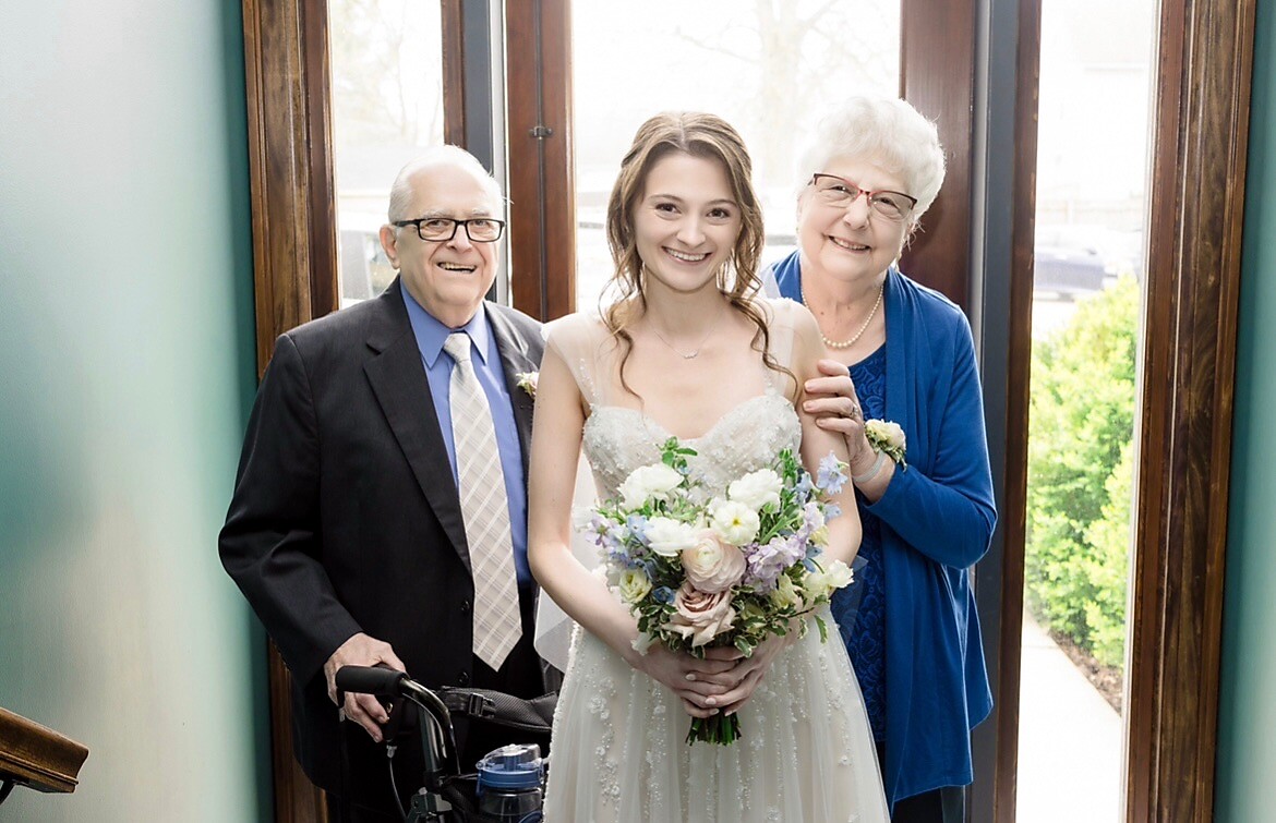 Bride in a beaded wedding gown holding a pastel bouquet, standing between her smiling elderly parents in a doorway.