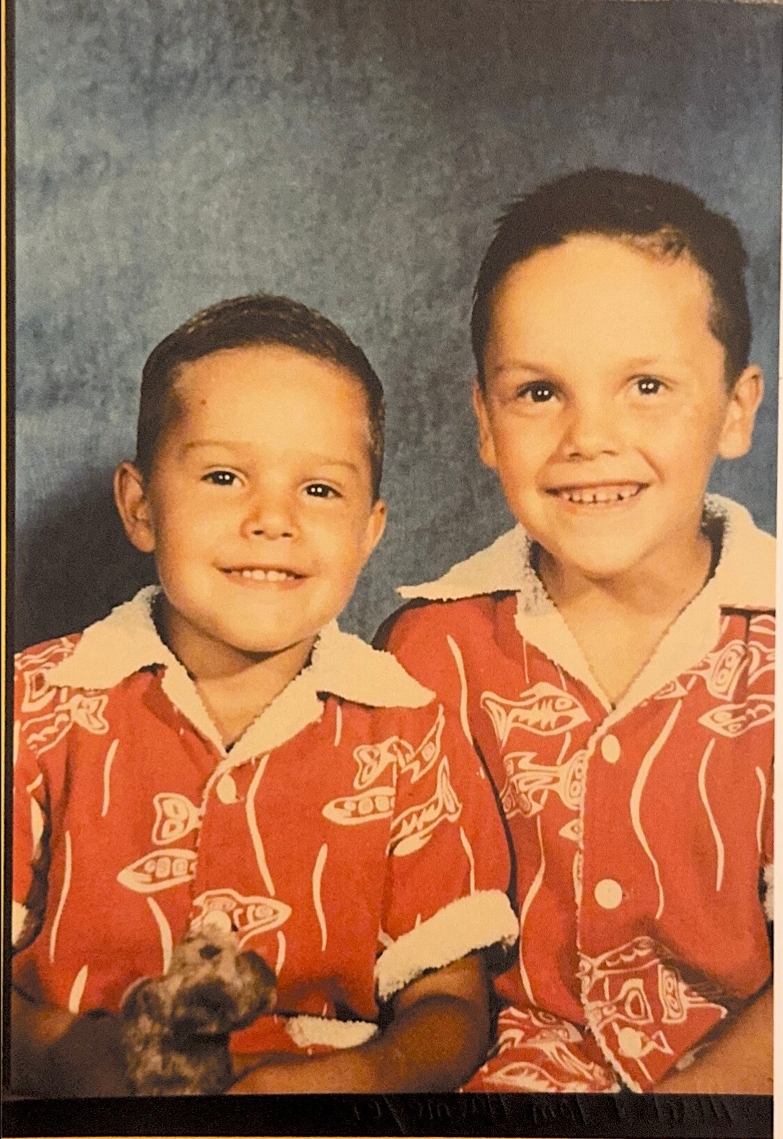 Two smiling young boys in matching red patterned shirts with white collars pose together against a blue backdrop, with a small dog visible at the bottom edge.