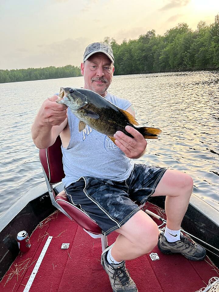 Man in a cap on a small boat holding up a largemouth bass he just caught, with a calm lake and tree line in the background.