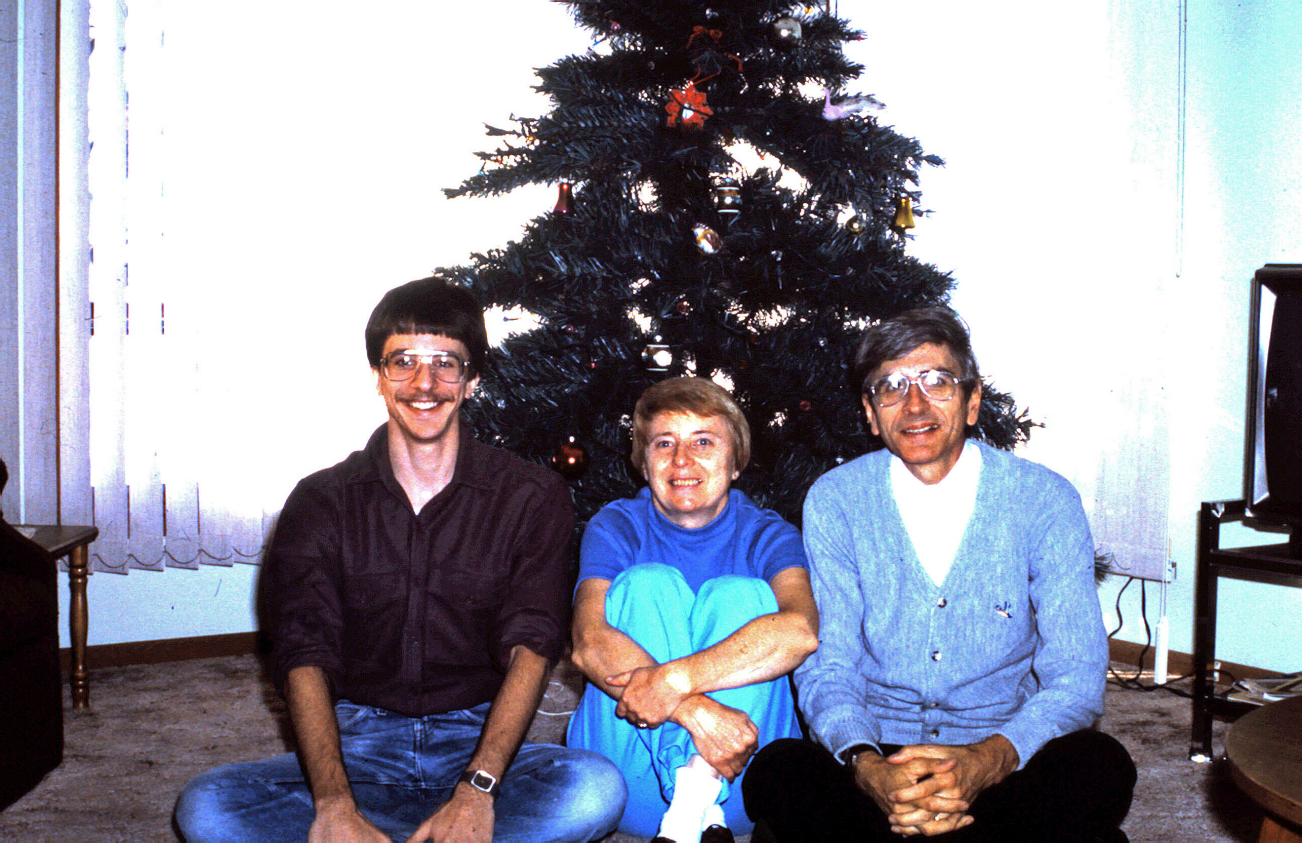 Three adults pose on the floor in front of a decorated Christmas tree indoors, smiling for a family photo.