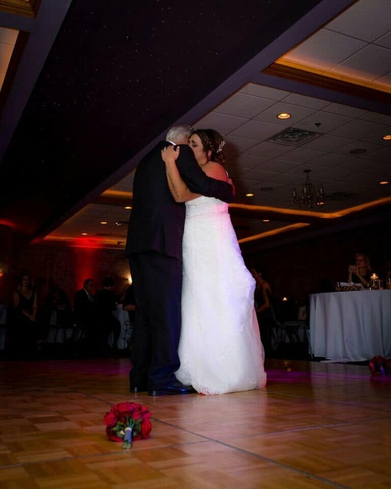 Bride in a white gown and a man in a dark suit share a close embrace while dancing in a dim ballroom; a red bouquet lies on the floor nearby.