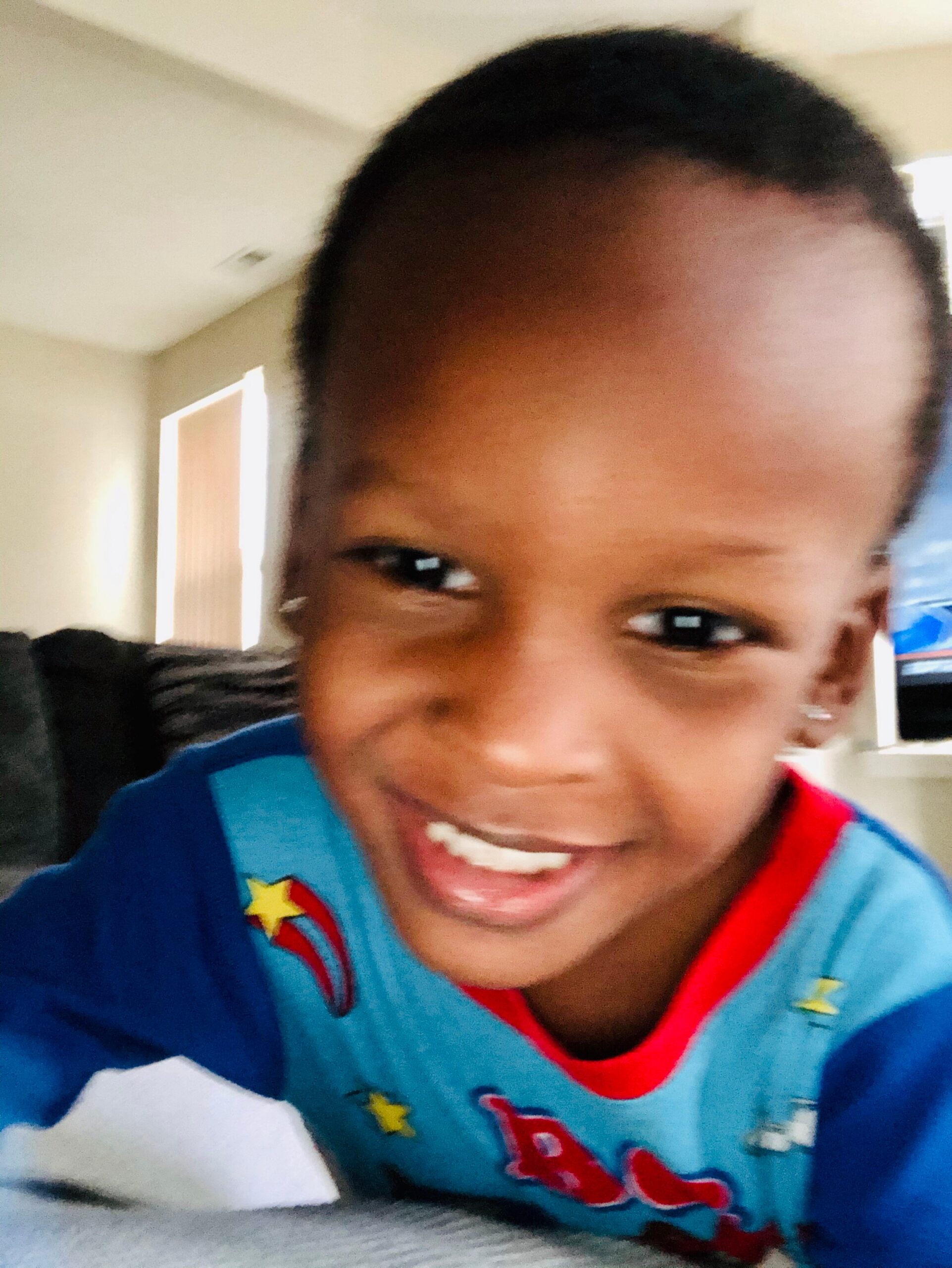 Close-up of a smiling child wearing a blue shirt with red trim and star patterns indoors, visible partial face.