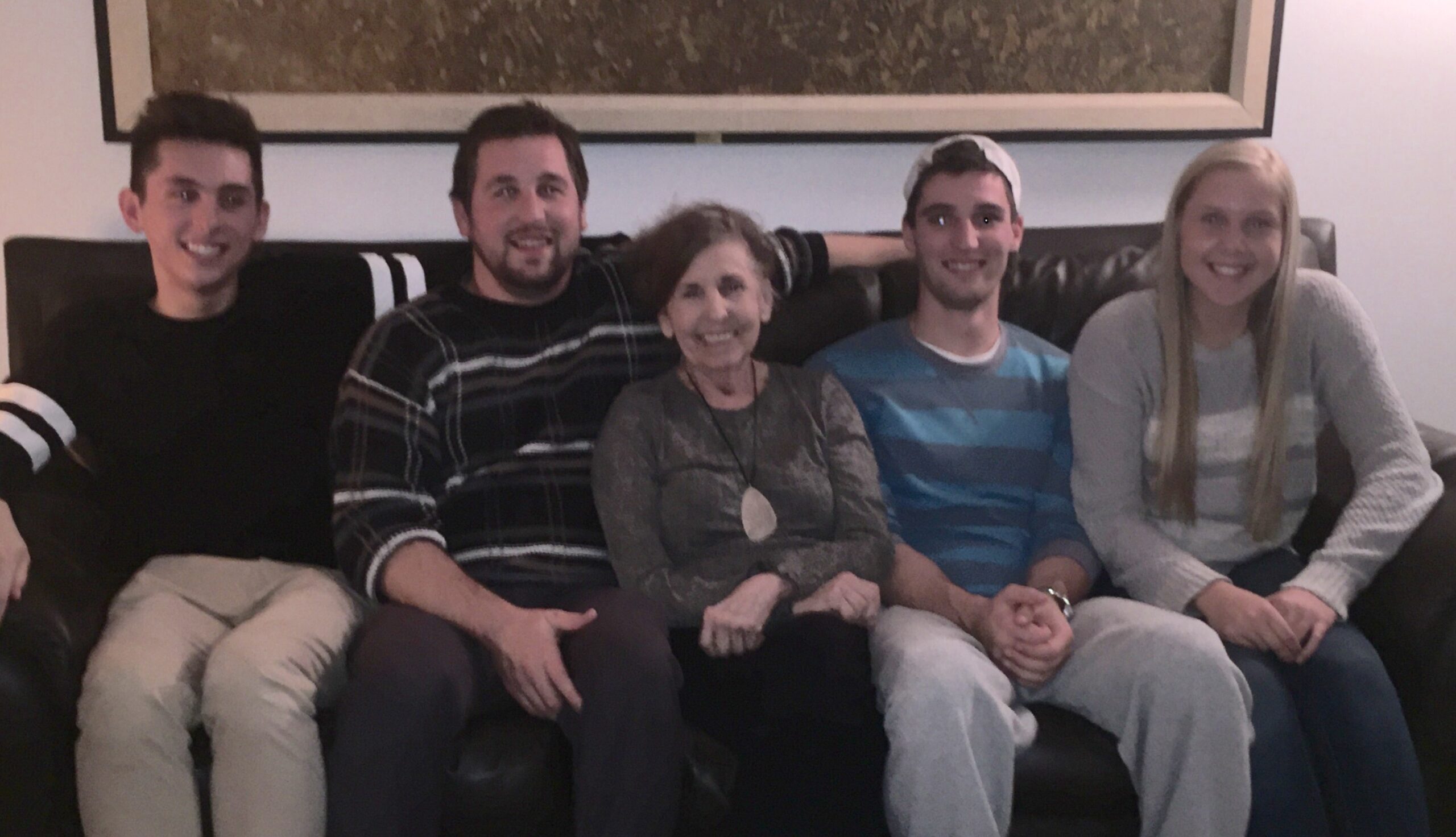 Five people sit closely on a dark sofa, smiling at the camera in a casual living-room setting.
