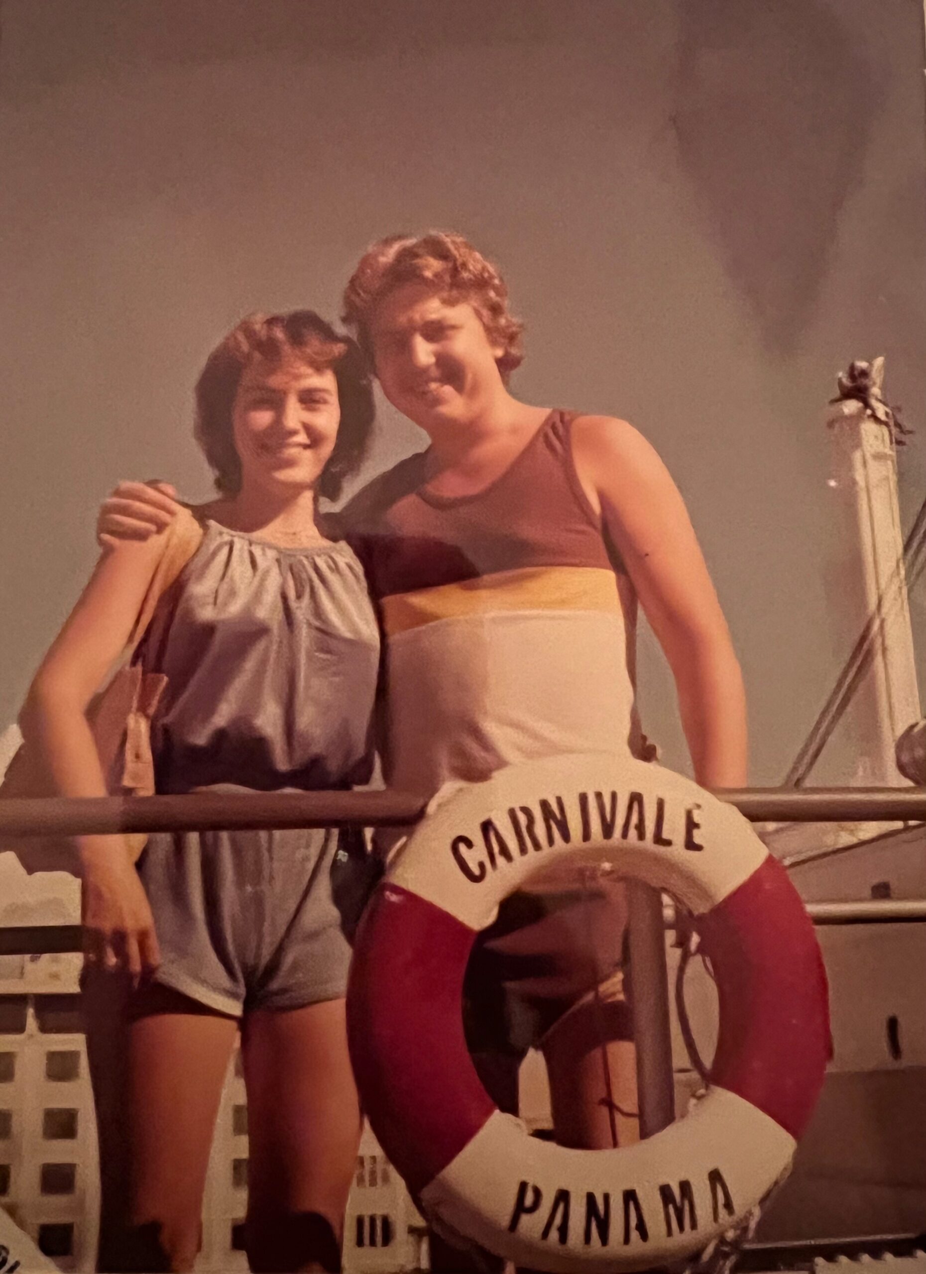 Two smiling friends pose together on a carnival ship railing, one in a blue dress, the other in a sleeveless top. A Panama life preserver hangs nearby.