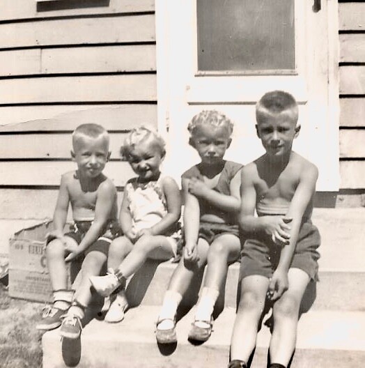 Four young children sit on the front steps of a wooden house; a girl in a dress sits second from the left among three boys.