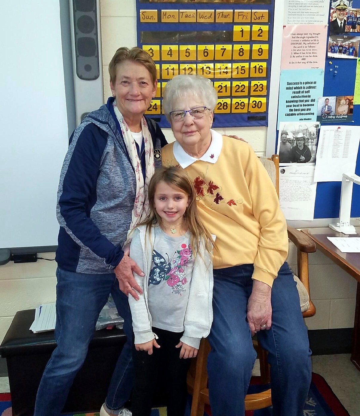 Three generations posing together in a classroom, smiling at the camera with a yellow calendar grid on the wall behind them.
