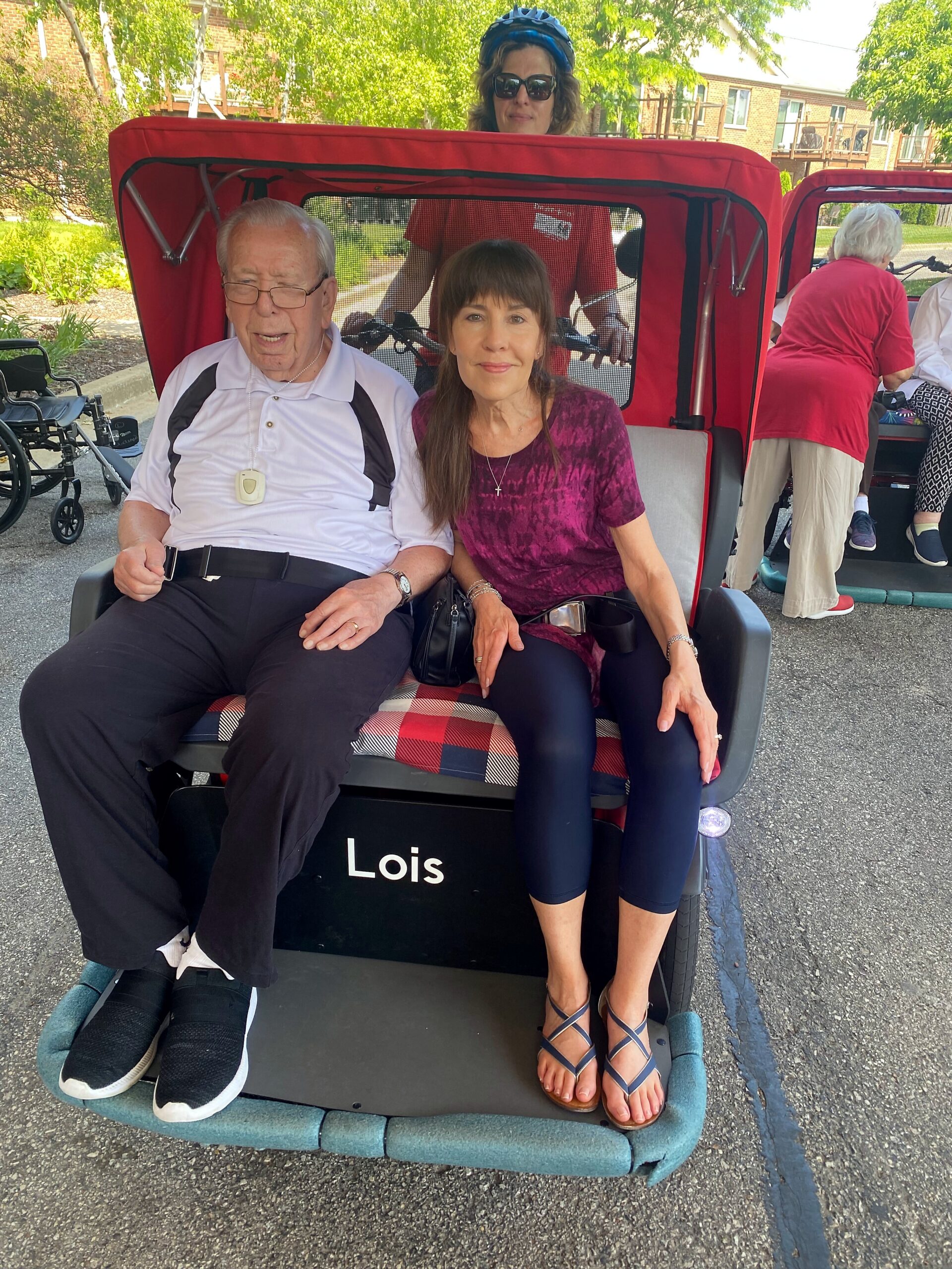 An elderly man and a woman sit together in a red-covered pedicab labeled 'Lois' outdoors, with helpers and others nearby in a sunny park setting.