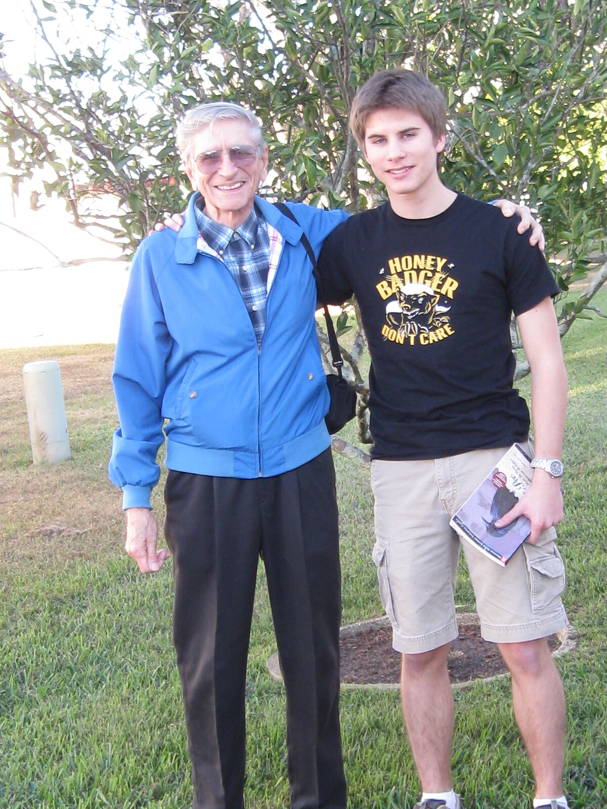 Two men pose together outdoors: an elderly man in a blue jacket and sunglasses with a plaid shirt, and a younger man in a black graphic t‑shirt, arms around each other in a grassy area with trees behind them.