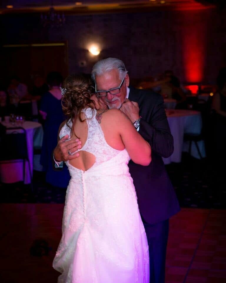 Older man in a dark suit dancing closely with a bride in a white lace gown at a reception hall.