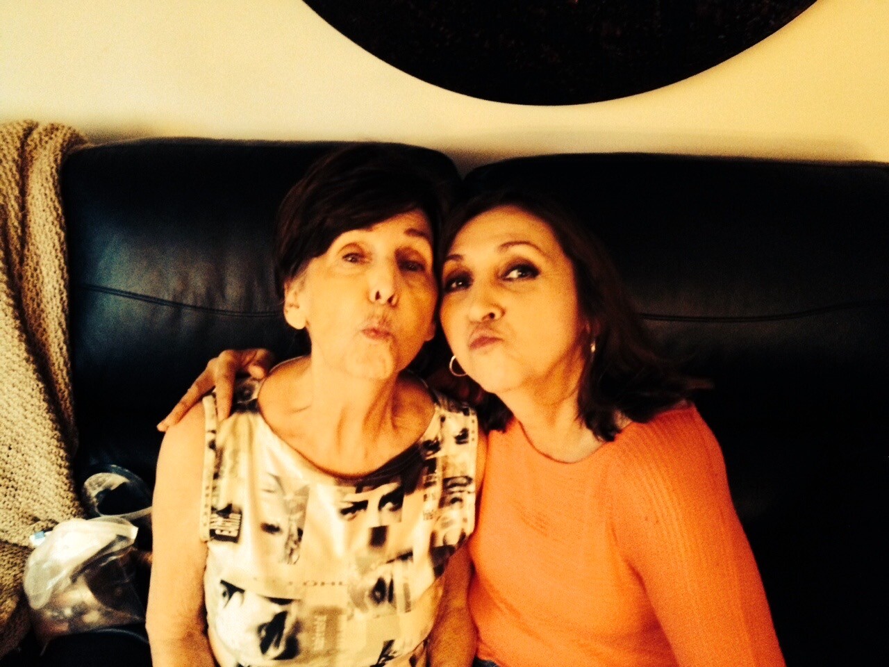 Two women sit closely on a dark couch, pursing their lips at the camera in a playful kissy pose indoors.