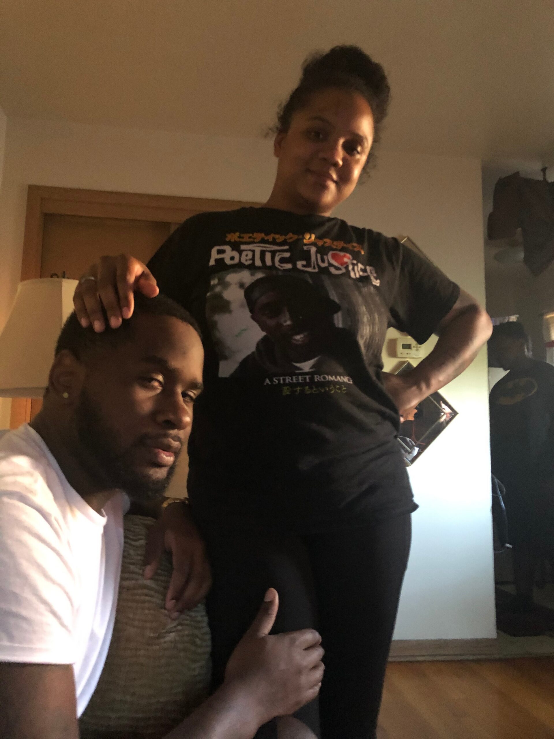 Two people indoors posing for a photo: a seated person in a white T-shirt and a standing person in a black graphic tee with their arm on the other's shoulder in a living room.