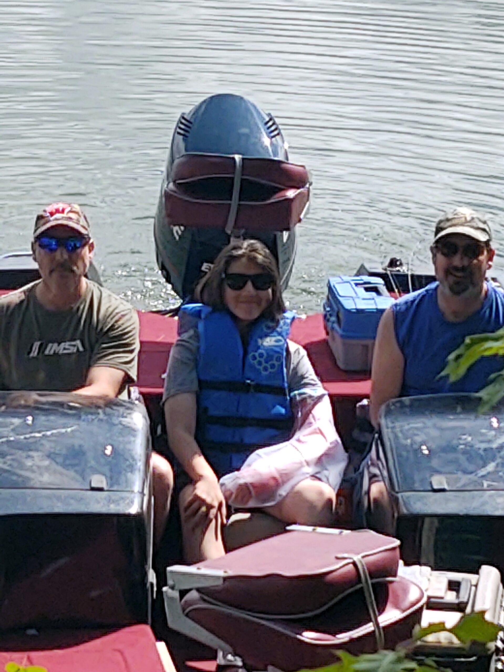 Three people sit in a small boat on calm water, wearing life jackets and sunglasses; a person in the middle smiles at the camera.