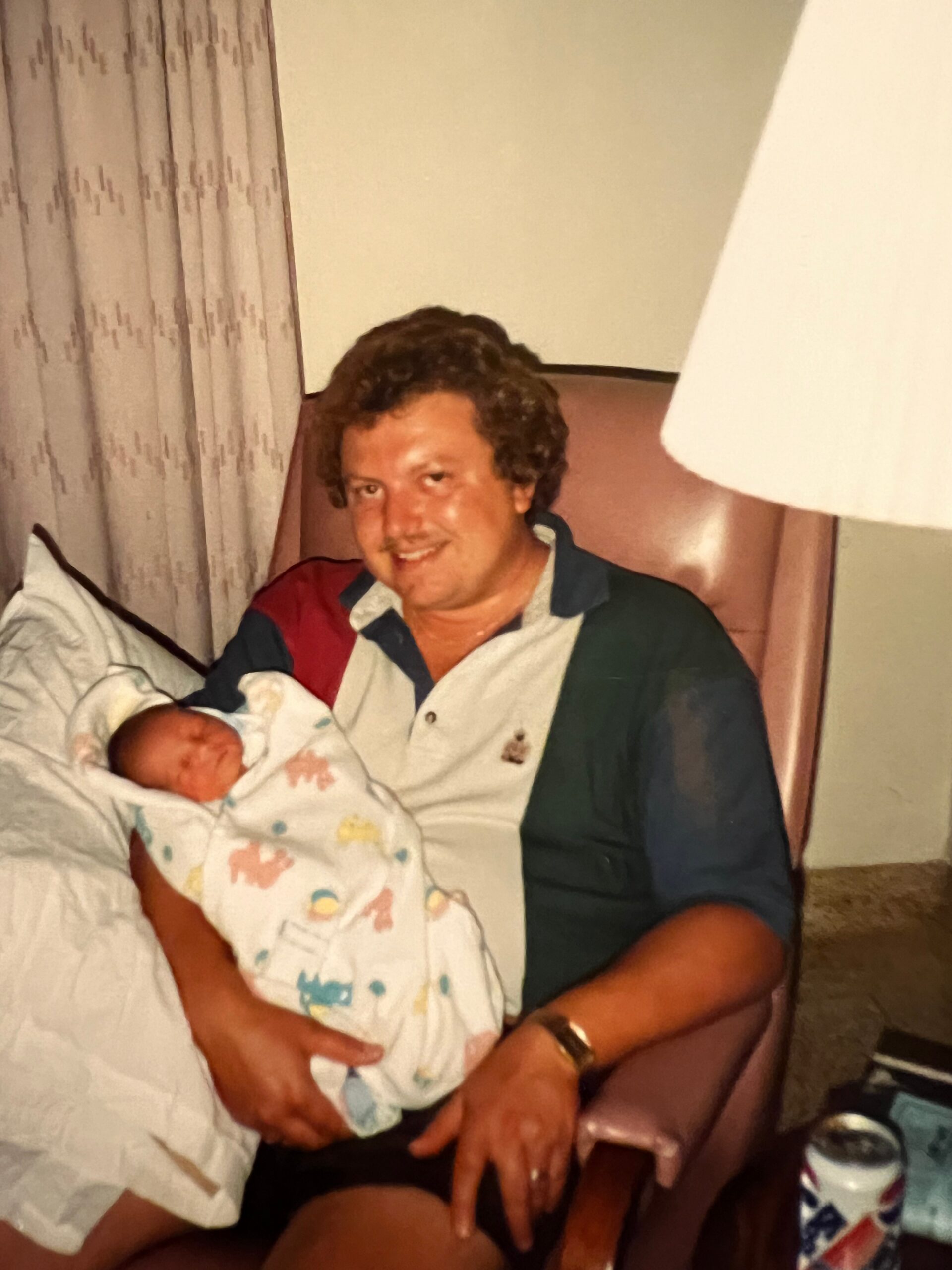 Man with a newborn baby wrapped in a pastel-printed blanket sits in a chair, smiling at the camera in a dimly lit room.