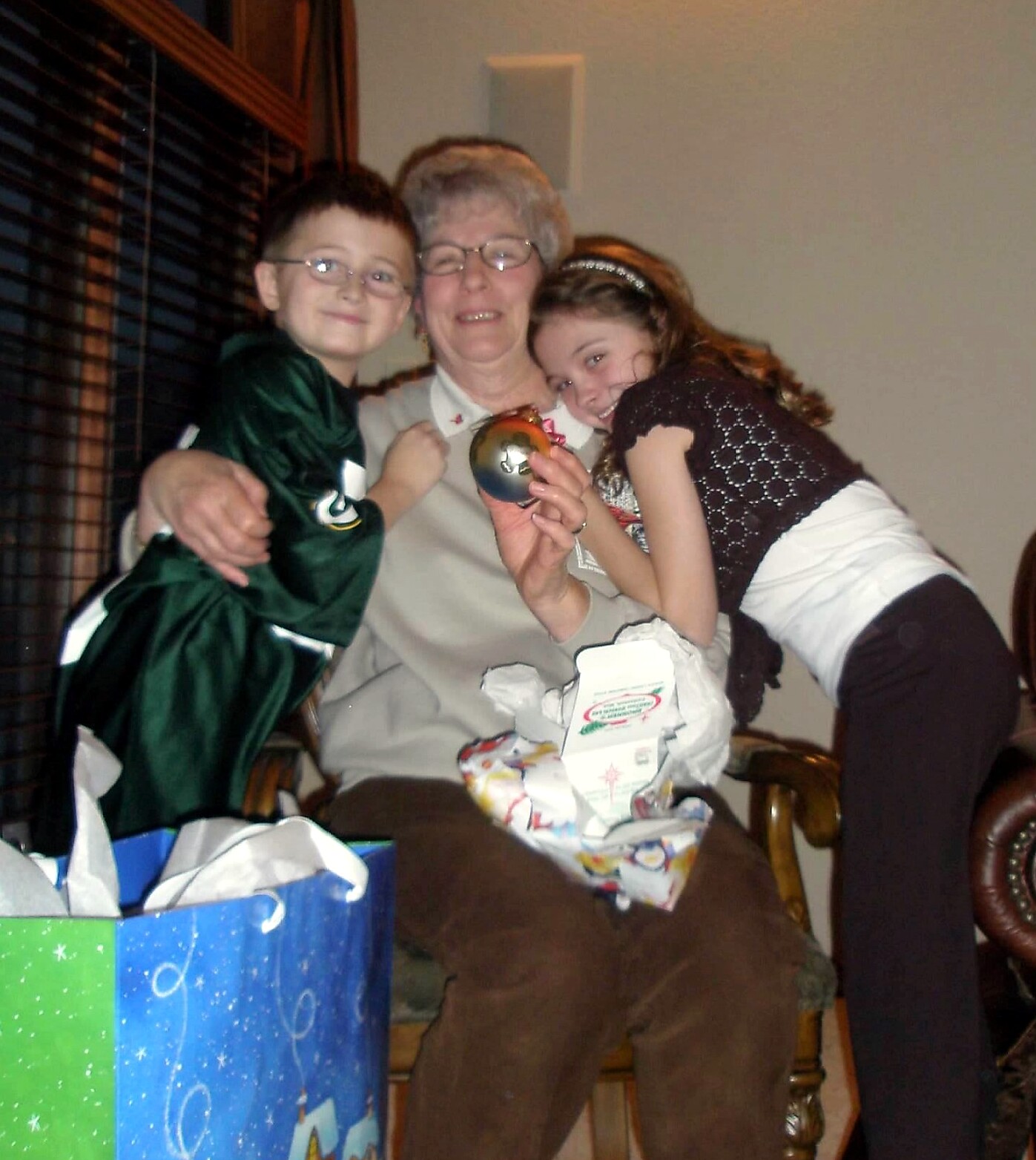 Grandmother sitting with two children, one boy in a green robe and a girl in a black top, all smiling as they pose with a metallic ornament amid wrapped gifts and gift bags on the floor.