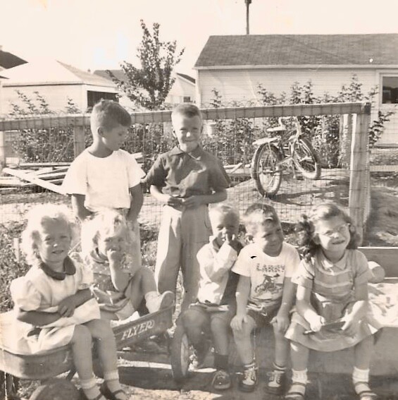 A group of seven young children pose on a wooden bench in a yard, with a bicycle and fence in the background.