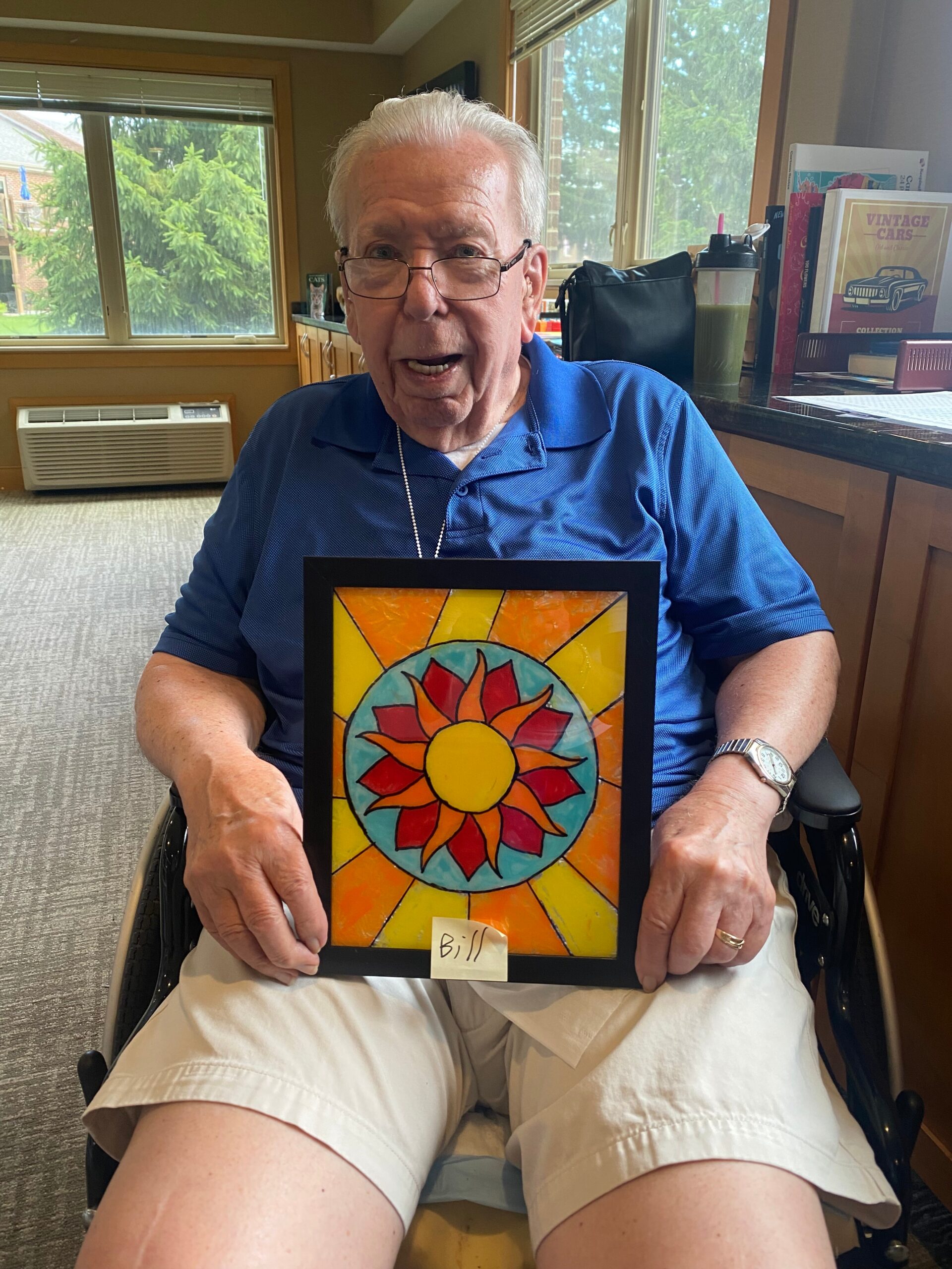 Older man in a wheelchair holds a colorful sunburst artwork framed in black; label reads 'Bill' at bottom edge, in a sunny indoor room.