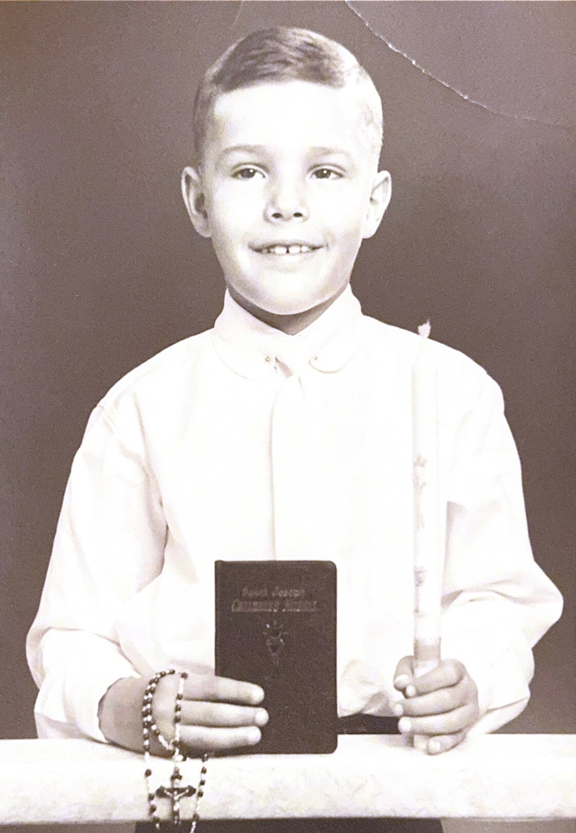 Black-and-white portrait of a smiling boy in a white shirt holding a small dark book and a rosary draped over his wrist.