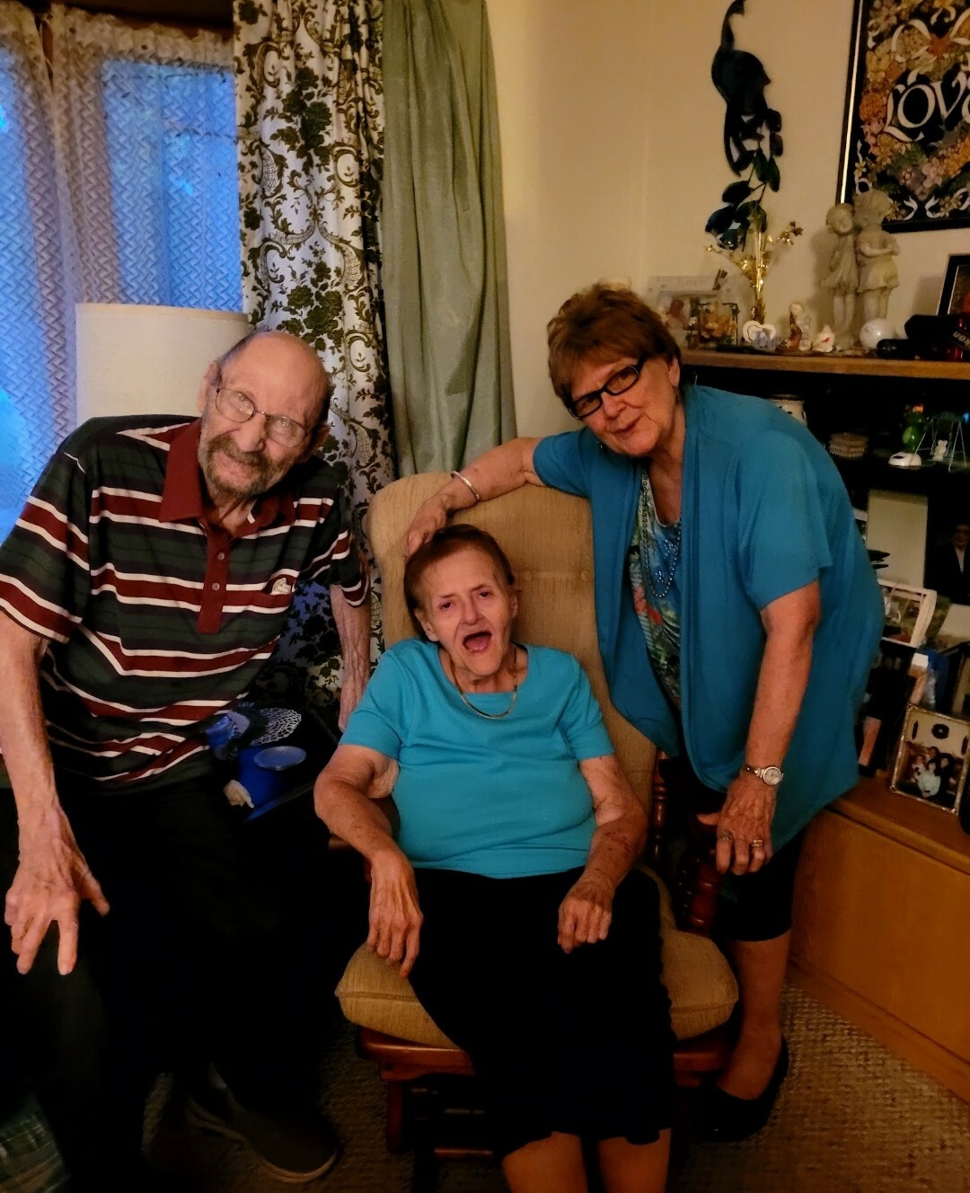 Three seniors posing in a cozy living room: a man on the left, a woman in a chair center, and another woman standing on the right with a hand on the chair.