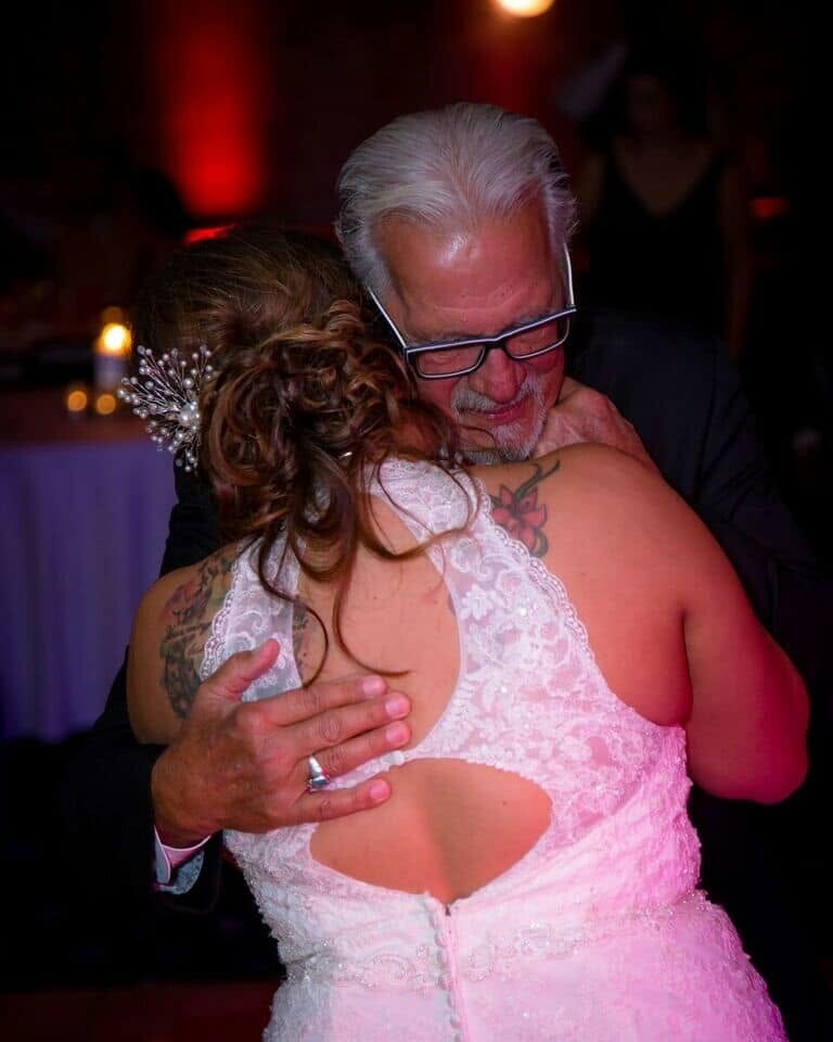 Older man with white hair hugs a bride in a lace dress, sharing a tender moment at a wedding dance, hands visible on her back.