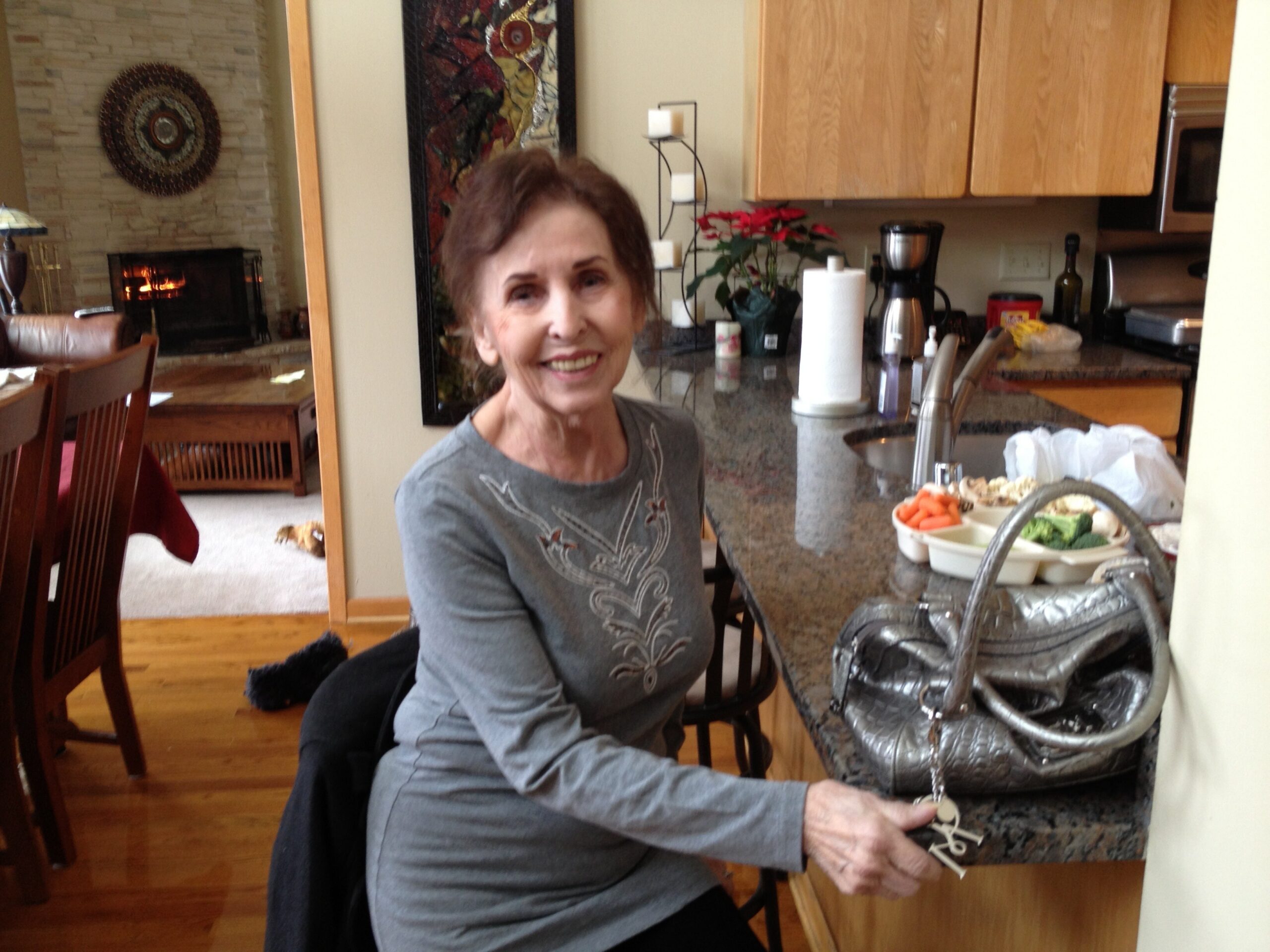 Smiling elderly woman in a gray shirt sits at a kitchen island, cutting with scissors near a basket of carrots and broccoli.
