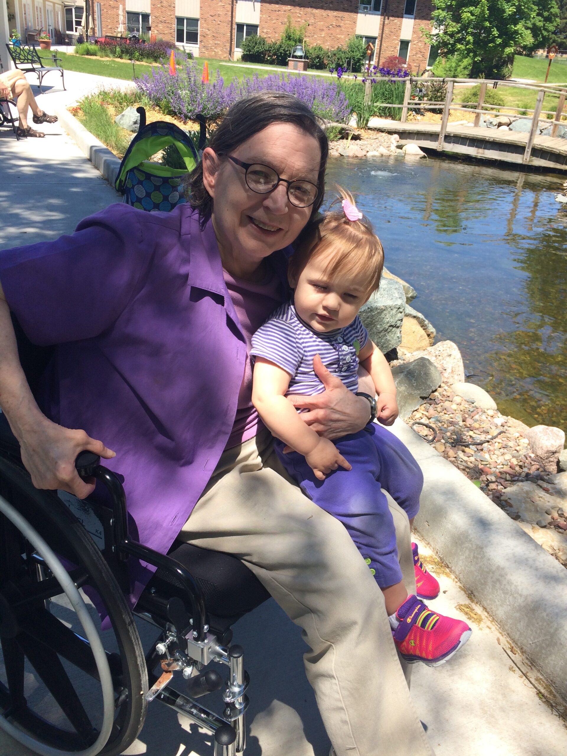 Woman in a purple shirt sits in a wheelchair, holding a young child by a pond in a sunny garden.