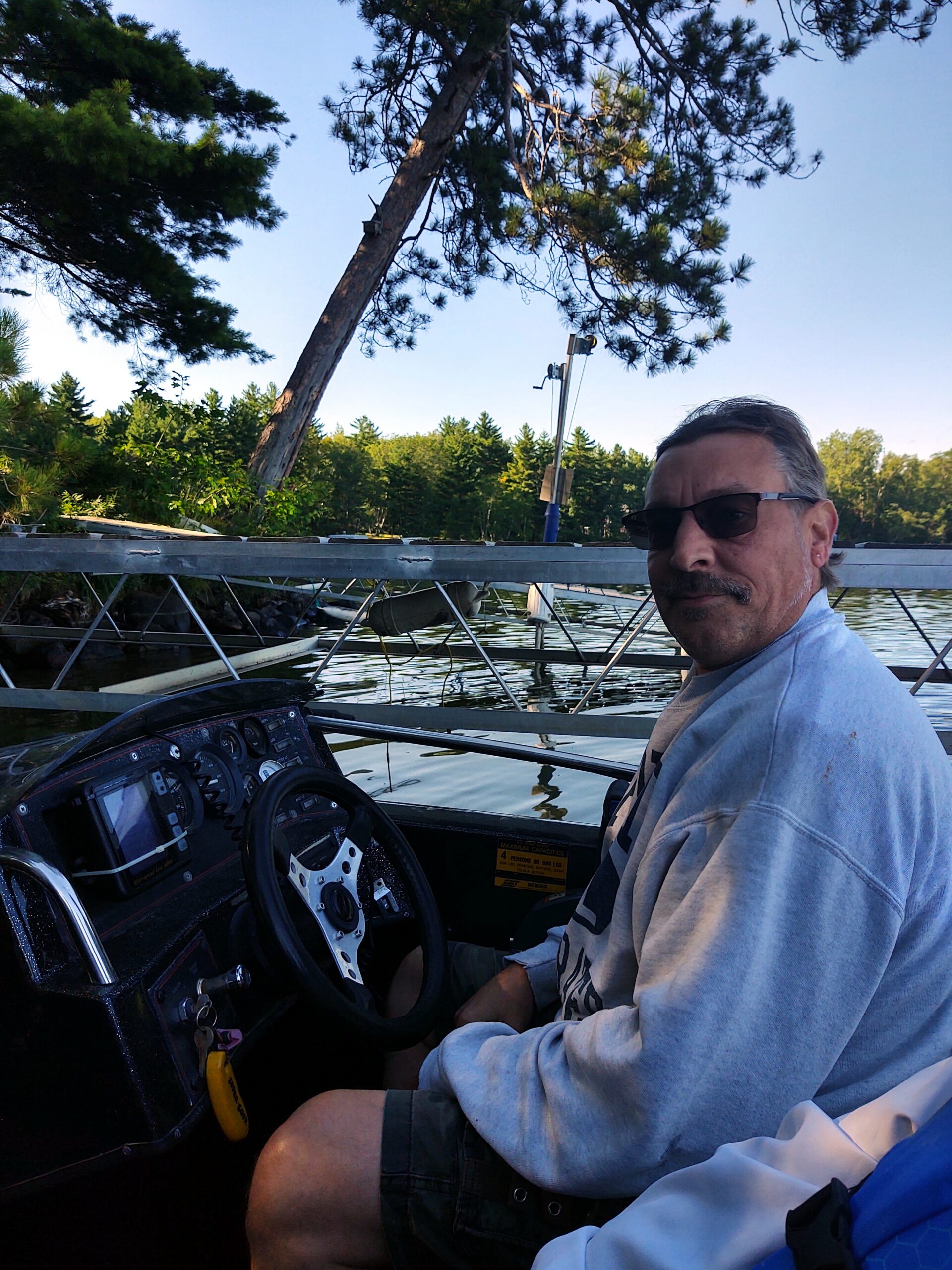 Man in a light sweatshirt sits at the boat helm, wearing sunglasses, on a calm lake with trees in the background.