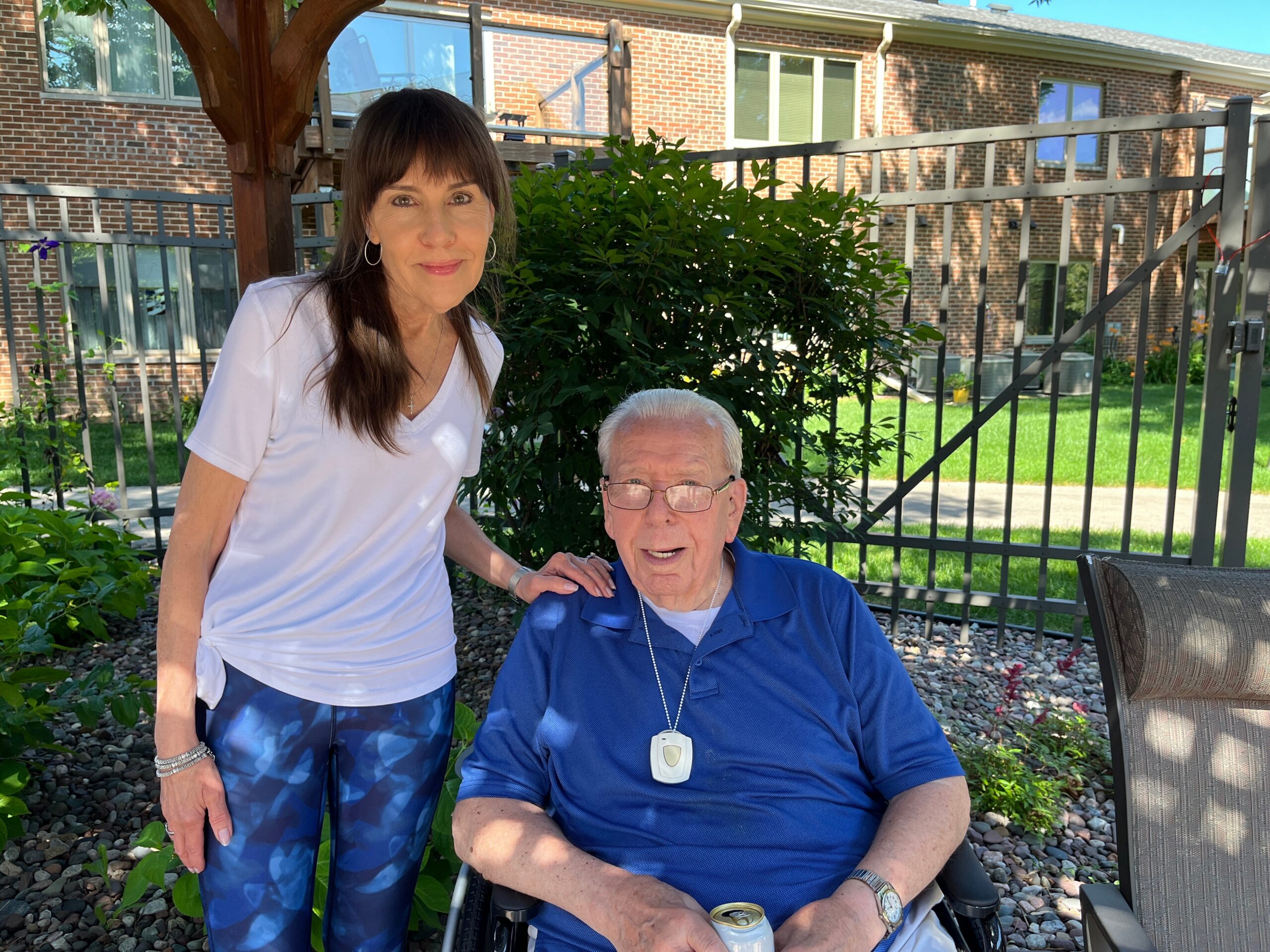 Smiling woman in a white top standing beside an elderly man in a wheelchair in a sunny backyard with a brick house and metal fence behind them.