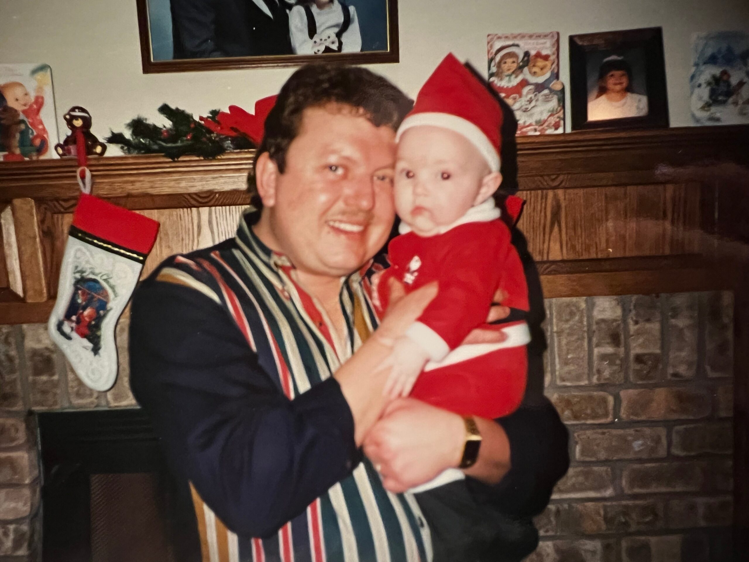 Man in a striped shirt holds a baby dressed in a red Santa suit by a brick fireplace with Christmas decor behind them.