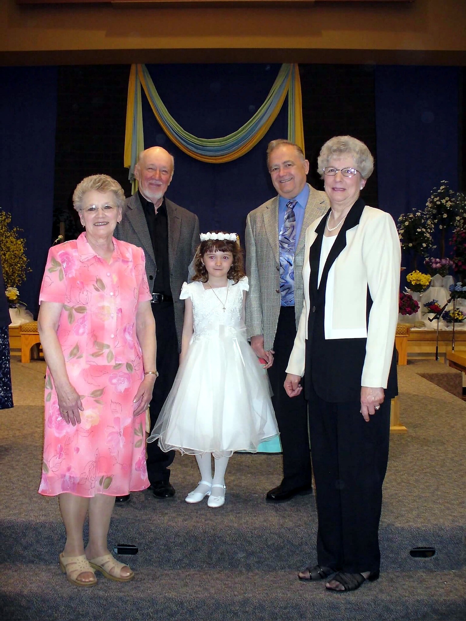 Group of five people posing on a stage in front of a blue and yellow curtain; a girl in a white dress with a flower crown stands center.