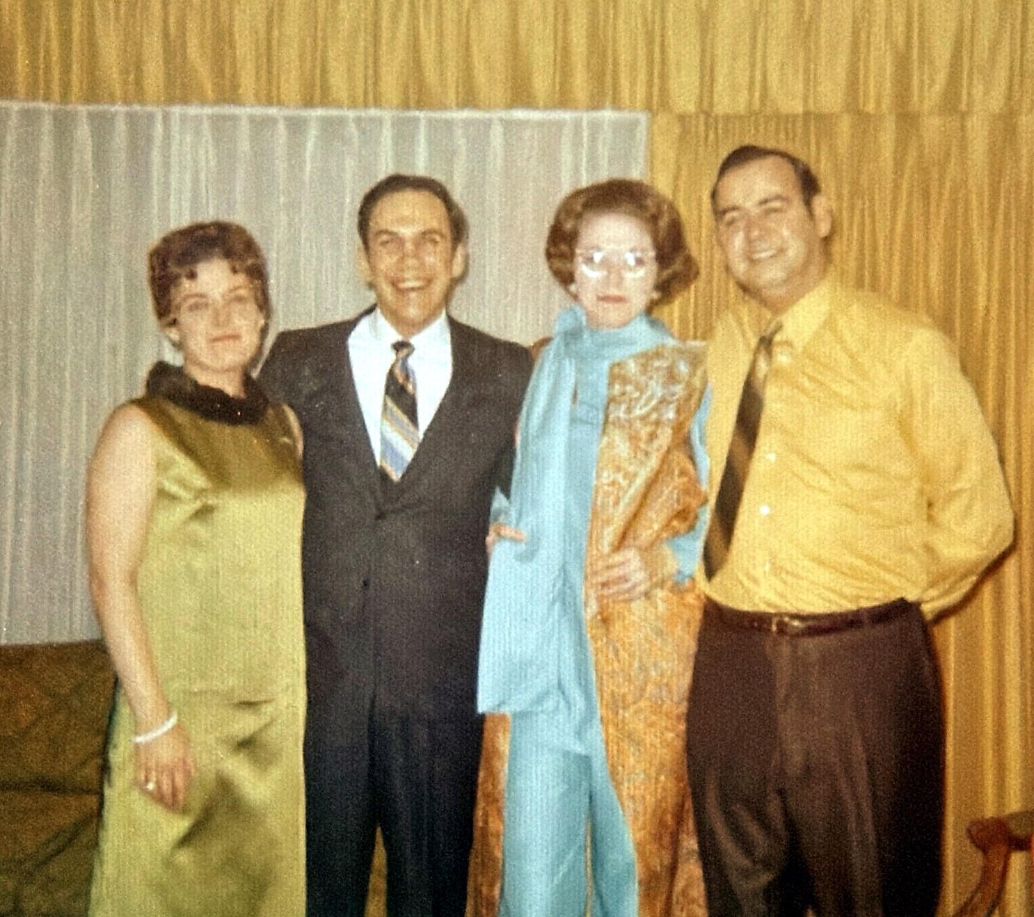 Four adults dressed in 1960s-style clothing pose together and smile for a group photo in a home setting.